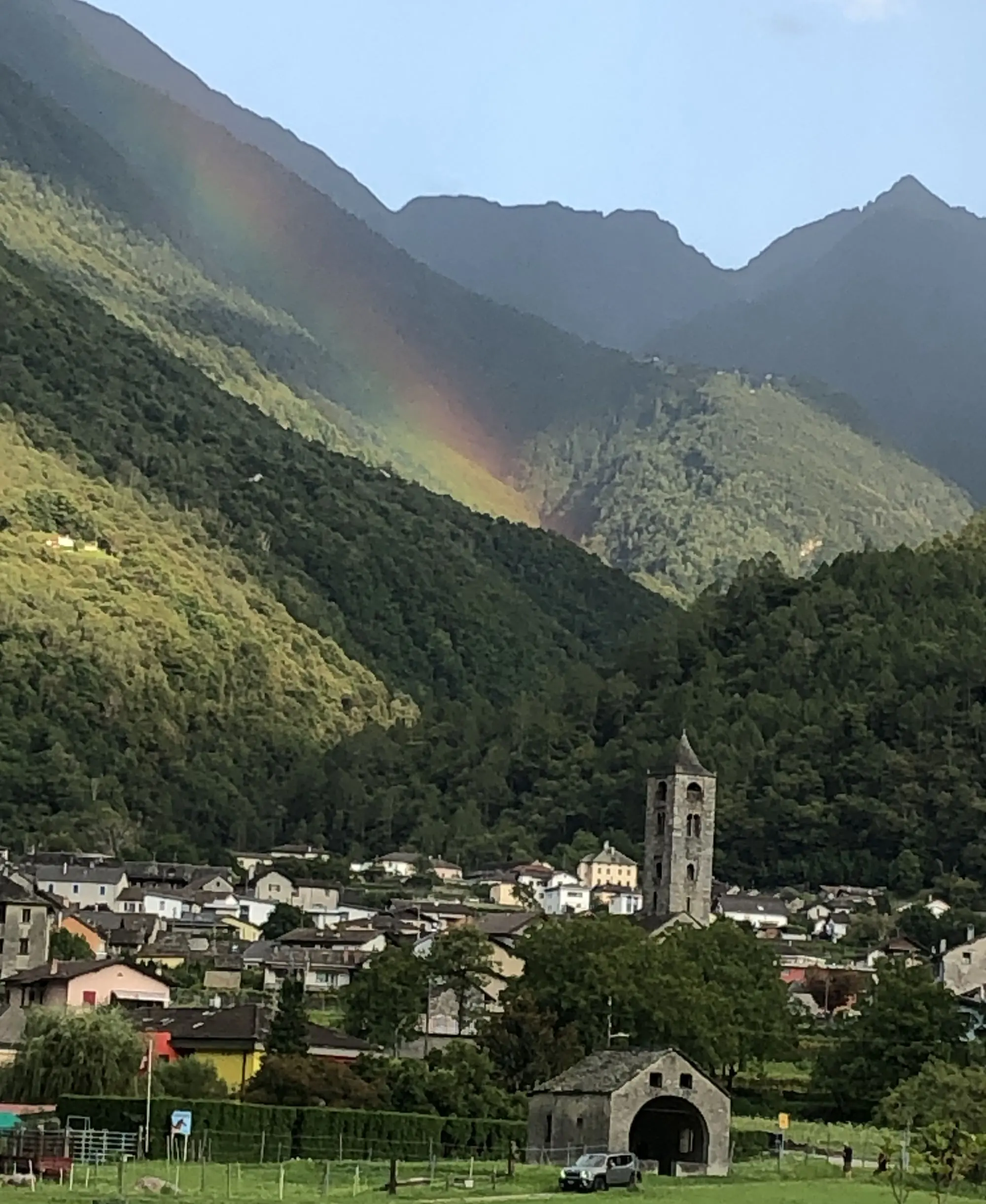 Panorama montano con un arcobaleno che si estende sopra le vette, un villaggio caratteristico con una chiesa e un campanile in primo piano.