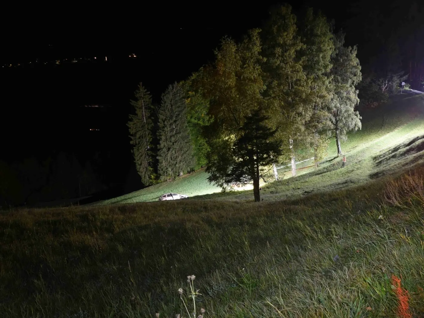 Un paesaggio notturno con una superficie erbosa illuminata da una luce artificiale. Alcuni alberi circondano l'area e in lontananza si intravedono delle luci. Una macchina è parcheggiata sulla collina.