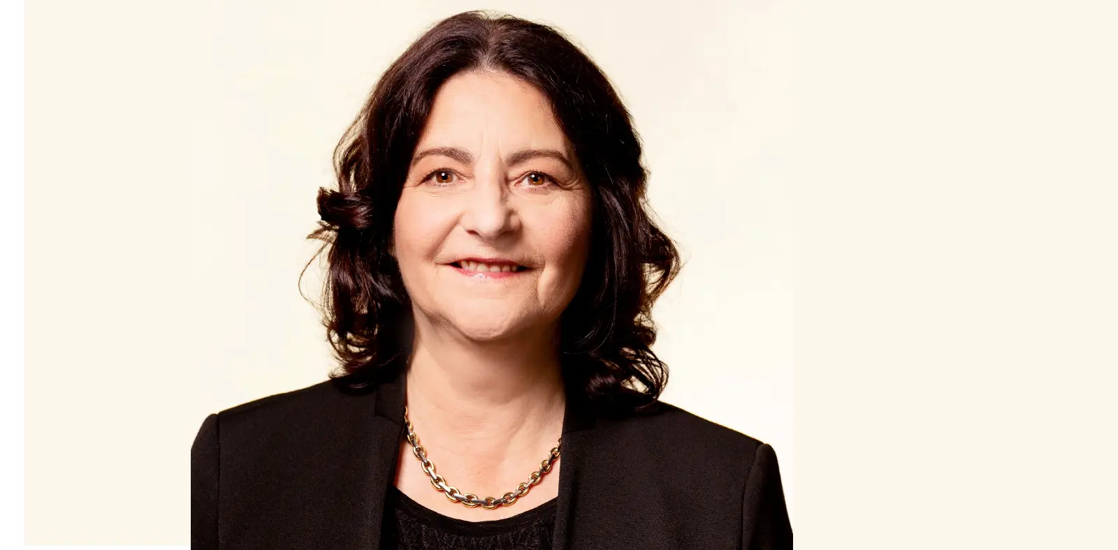 Una donna sorridente con capelli scuri e ricci, indossa un blazer nero e una collana. Lo sfondo è chiaro e neutro.