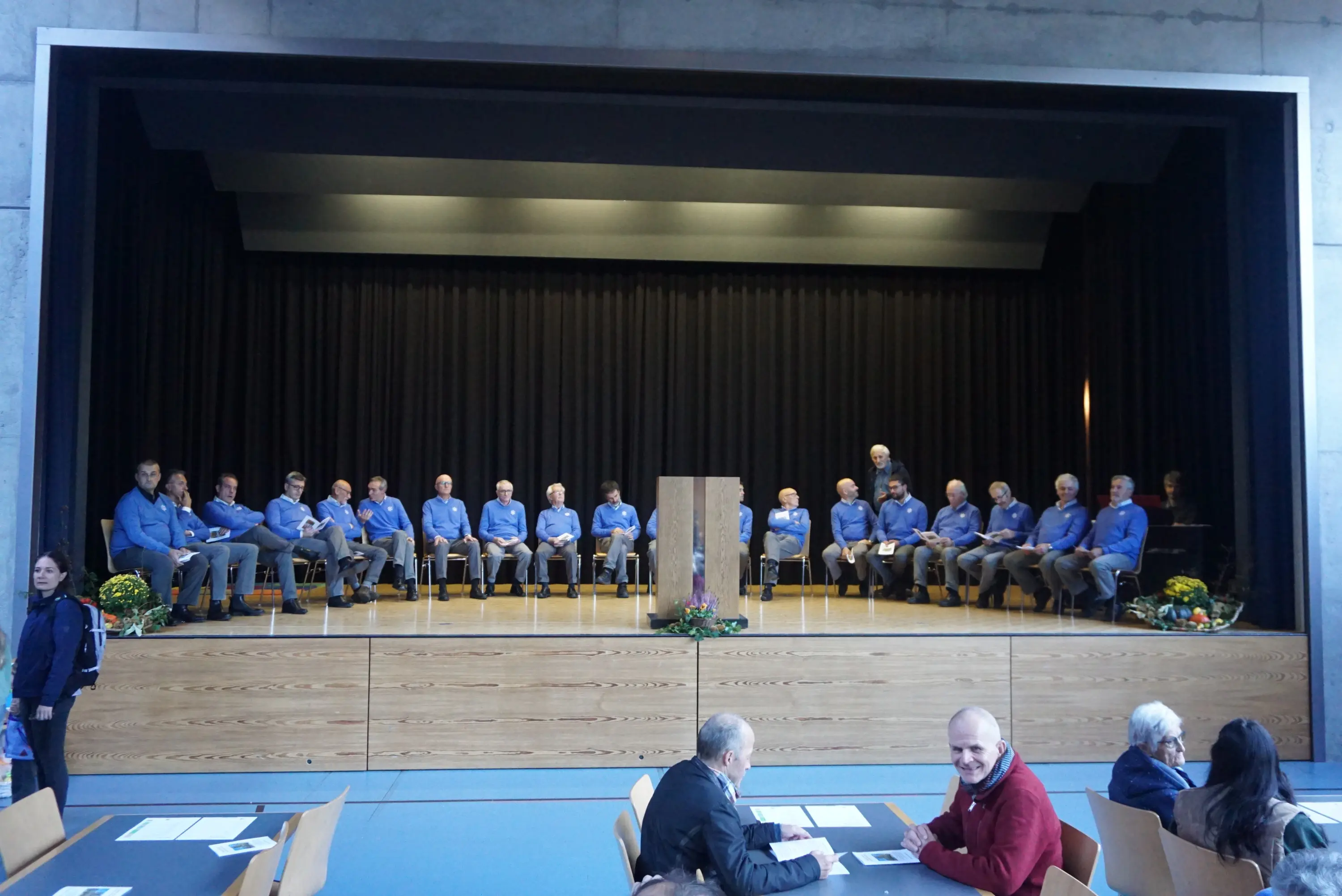 Un gruppo di persone sedute su un palco, vestite con camicie blu, in attesa di un evento. Alcuni membri sono in piedi accanto a un leggio, mentre il pubblico è visibile in primo piano.