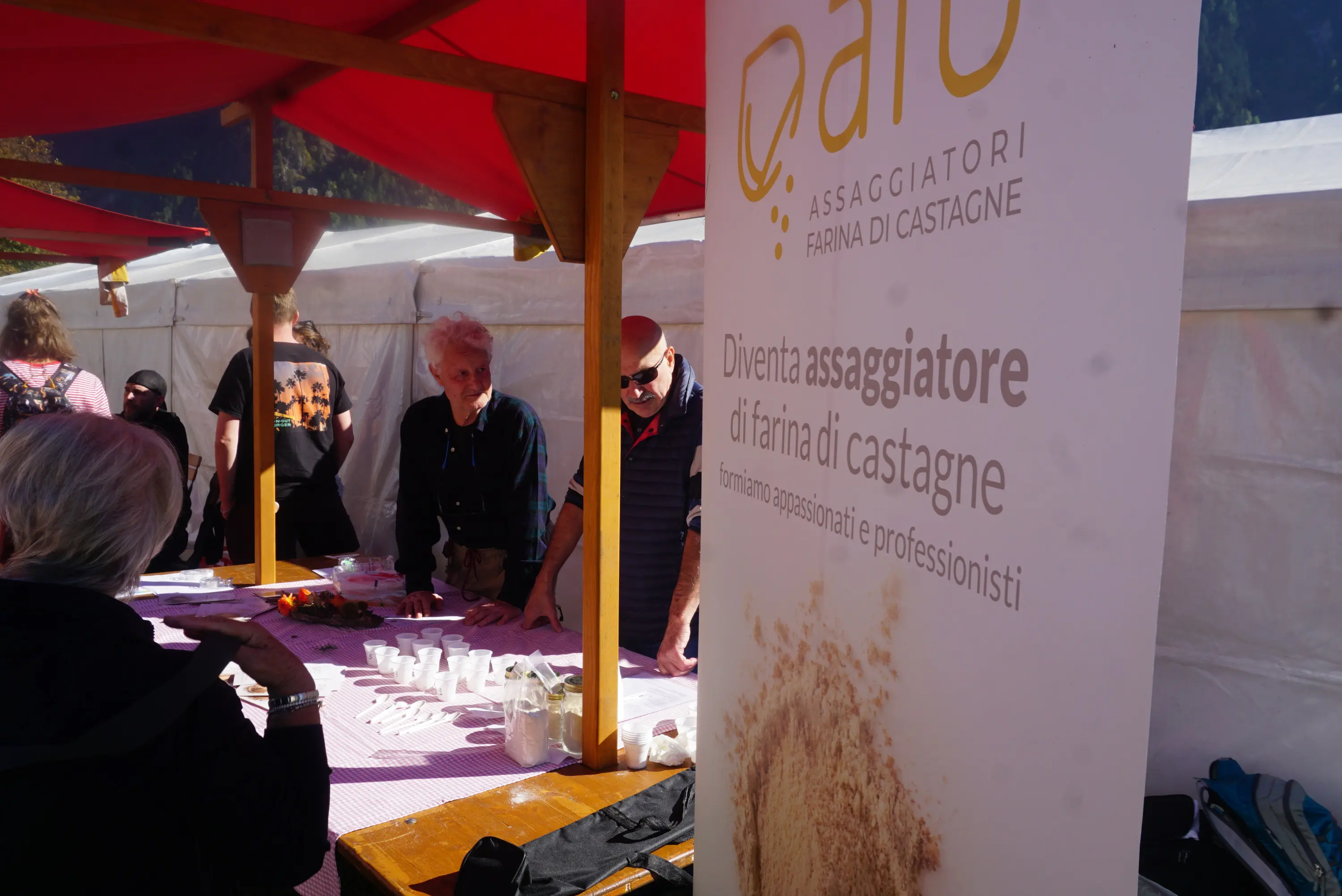 Un bancone al mercato con un'insegna che promuove la farina di castagne. Persone sono intente a lavorare e interagire. Si vedono materiali e utensili per la preparazione. Un'atmosfera festosa e accogliente.