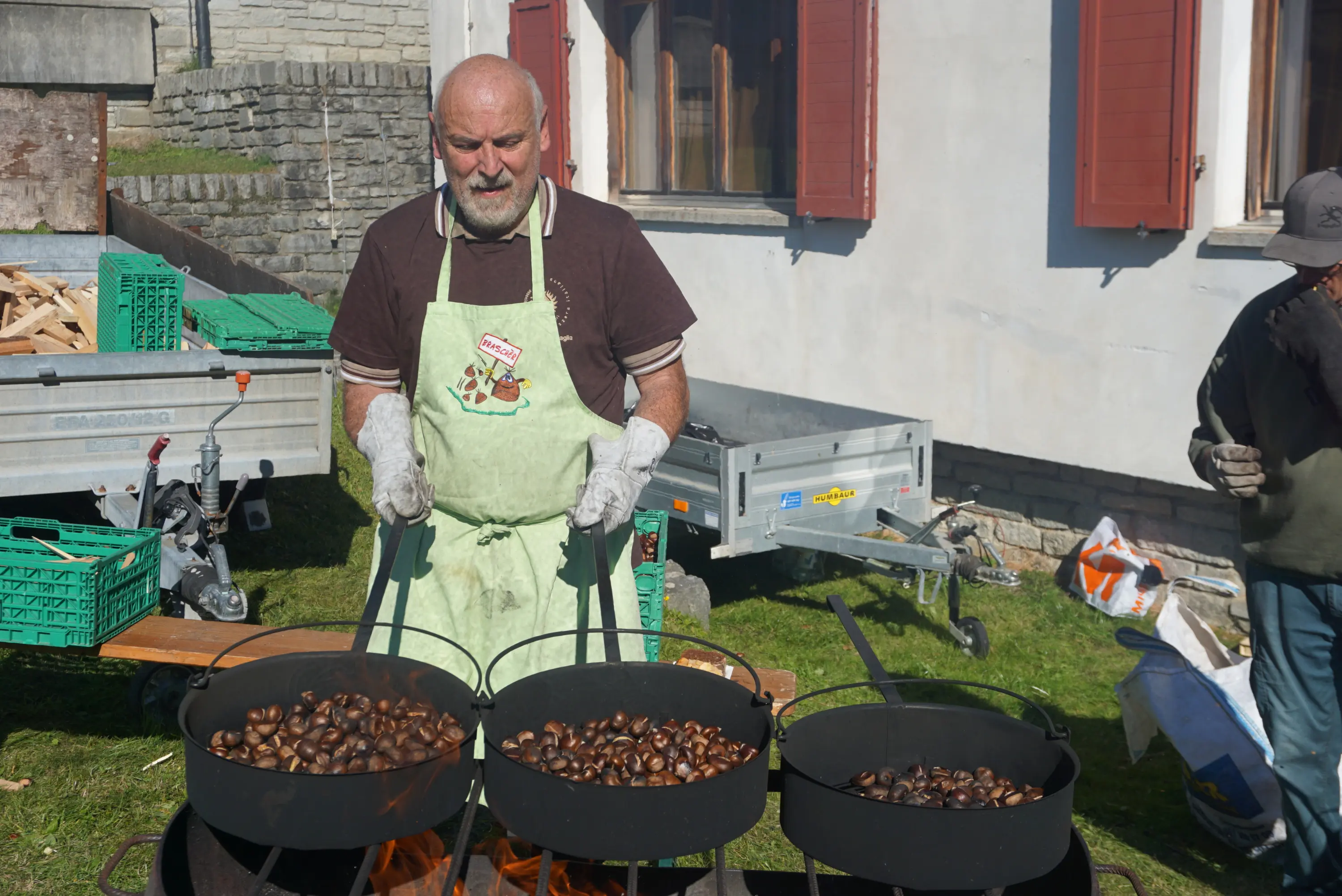 Un uomo con un grembiule verde sta cuocendo castagne in due pentole su un fuoco all'aperto. Sullo sfondo si vedono alcune strutture e cassette di raccolta.