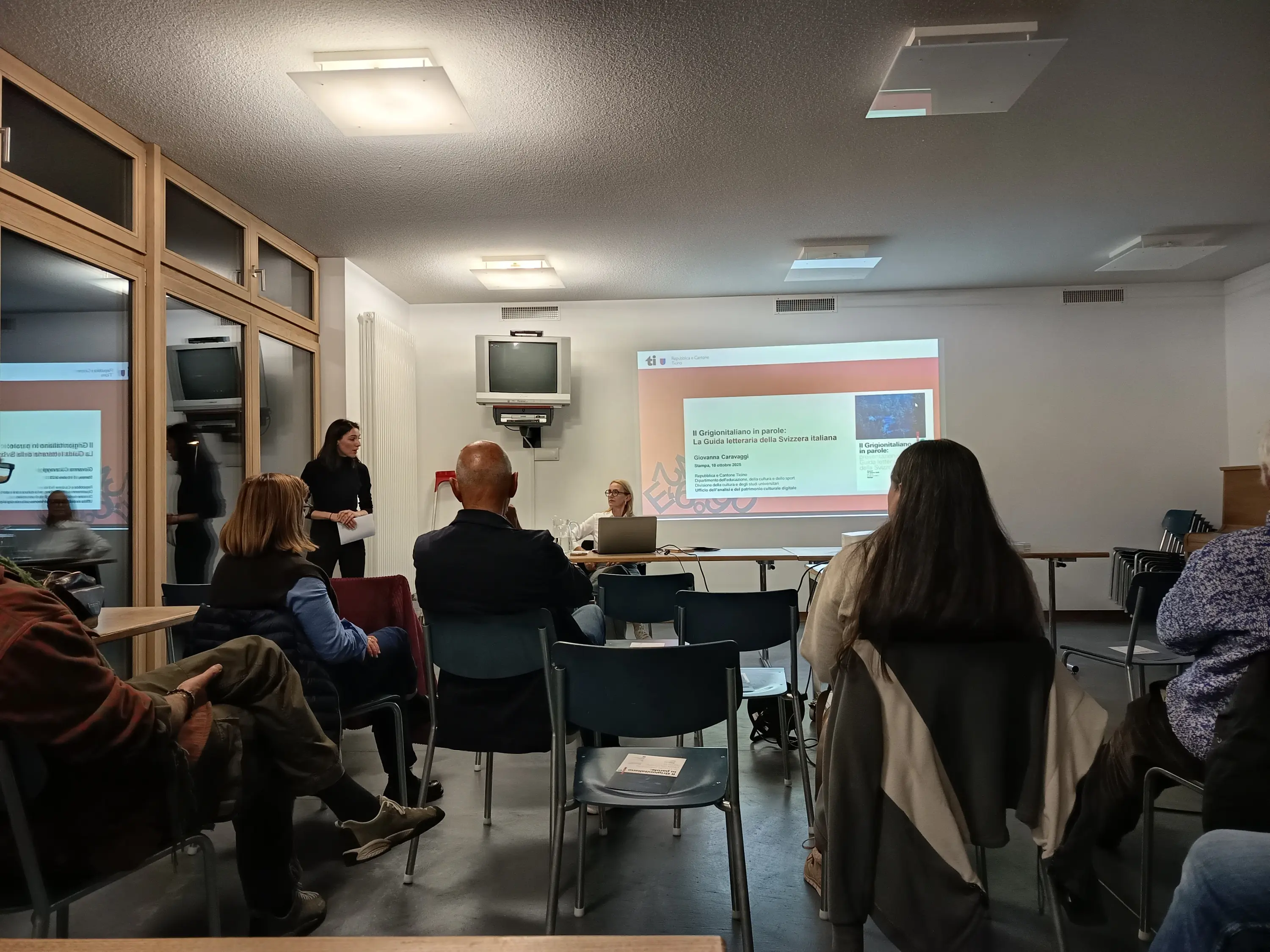 Una presentazione in corso in una sala riunioni. Una donna parla in piedi di fronte a un pubblico seduto, mentre un'altra donna è seduta a un tavolo con un computer. Sullo schermo è visibile una presentazione con testo.
