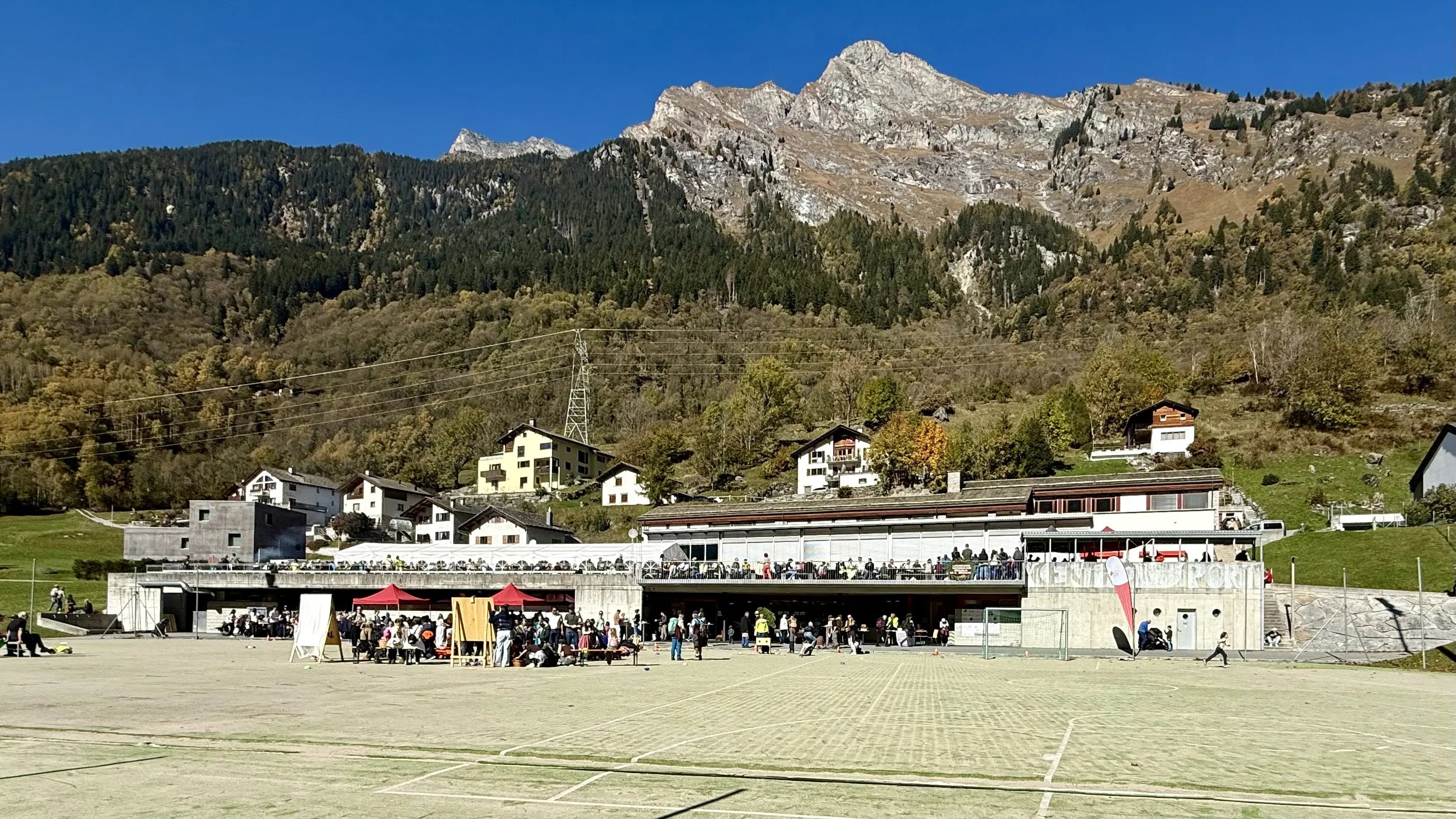 Una vista di un campo sportivo affollato con una tribuna e alcune strutture in un contesto montano. Sullo sfondo, si vedono alberi e montagne illuminate dal sole in una giornata serena. Diversi gruppi di persone sono presenti, e ci sono gazebo colorati.