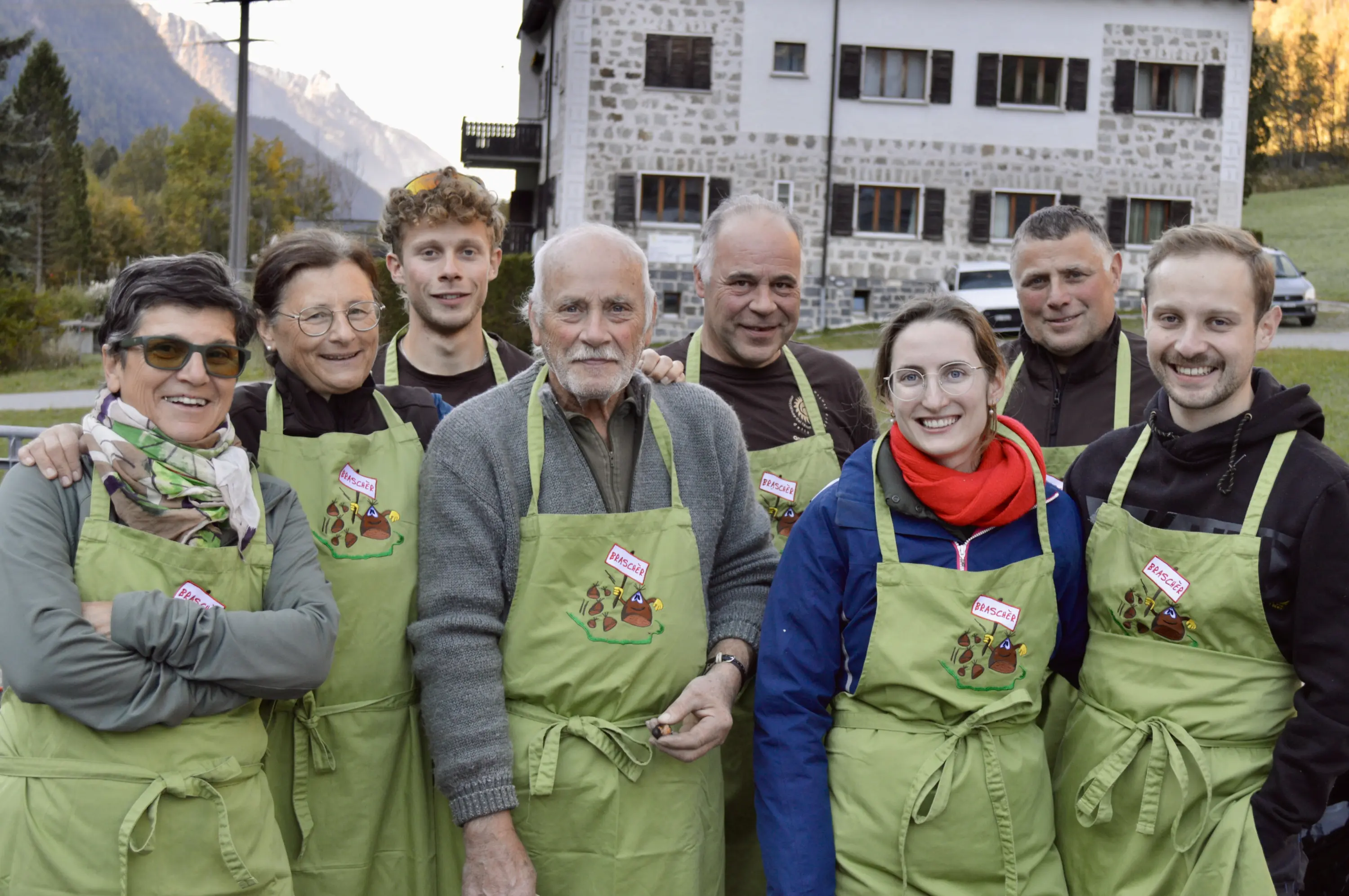 Un gruppo di otto persone sorridenti indossa grembiuli verdi. Si trovano all'aperto, con una casa in pietra sullo sfondo e montagne in lontananza. Tutti sembrano felici e pronti per un'attività culinaria.