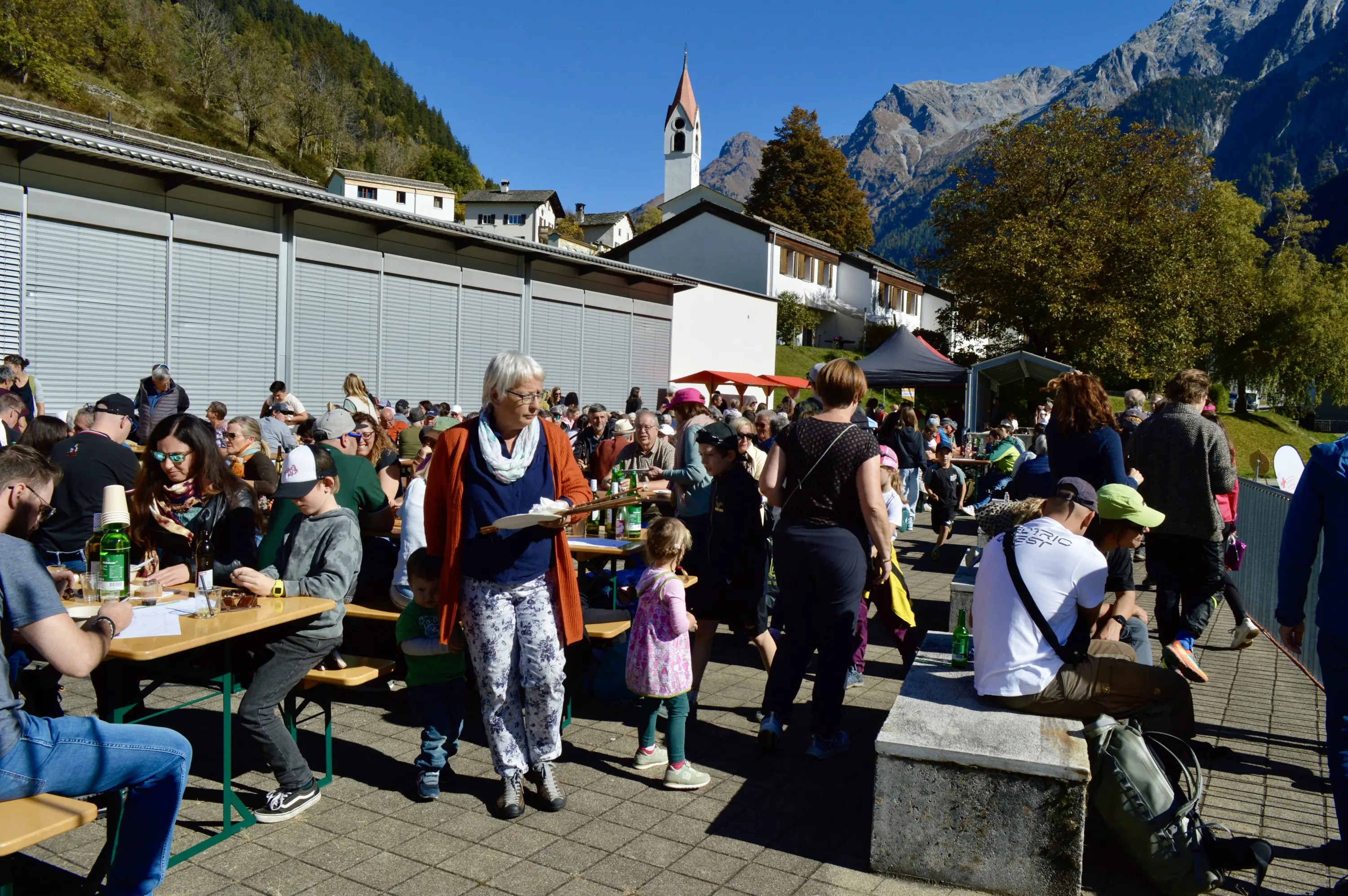 Una folla di persone in un'area all'aperto, sedute e in piedi attorno a tavoli, mentre mangiano e bevono. Alcuni bambini giocano tra gli adulti. Sullo sfondo si vedono montagne e un campanile. È una giornata di sole.