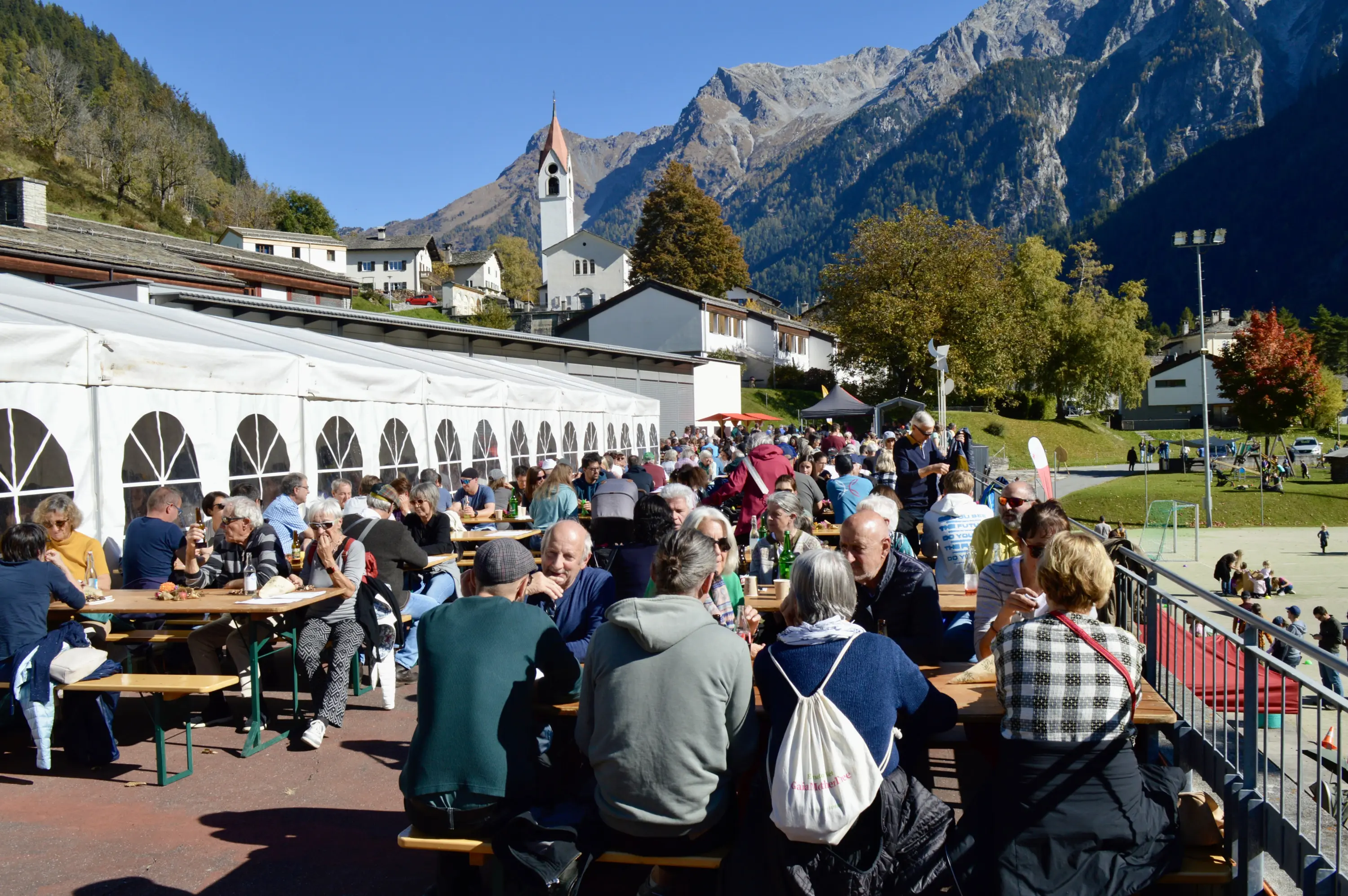 Una folla di persone sedute attorno a tavoli di legno all'aperto, mentre godono di un evento sociale. Sullo sfondo si vedono monti maestosi e una chiesa con un campanile. L'atmosfera è vivace e solare.