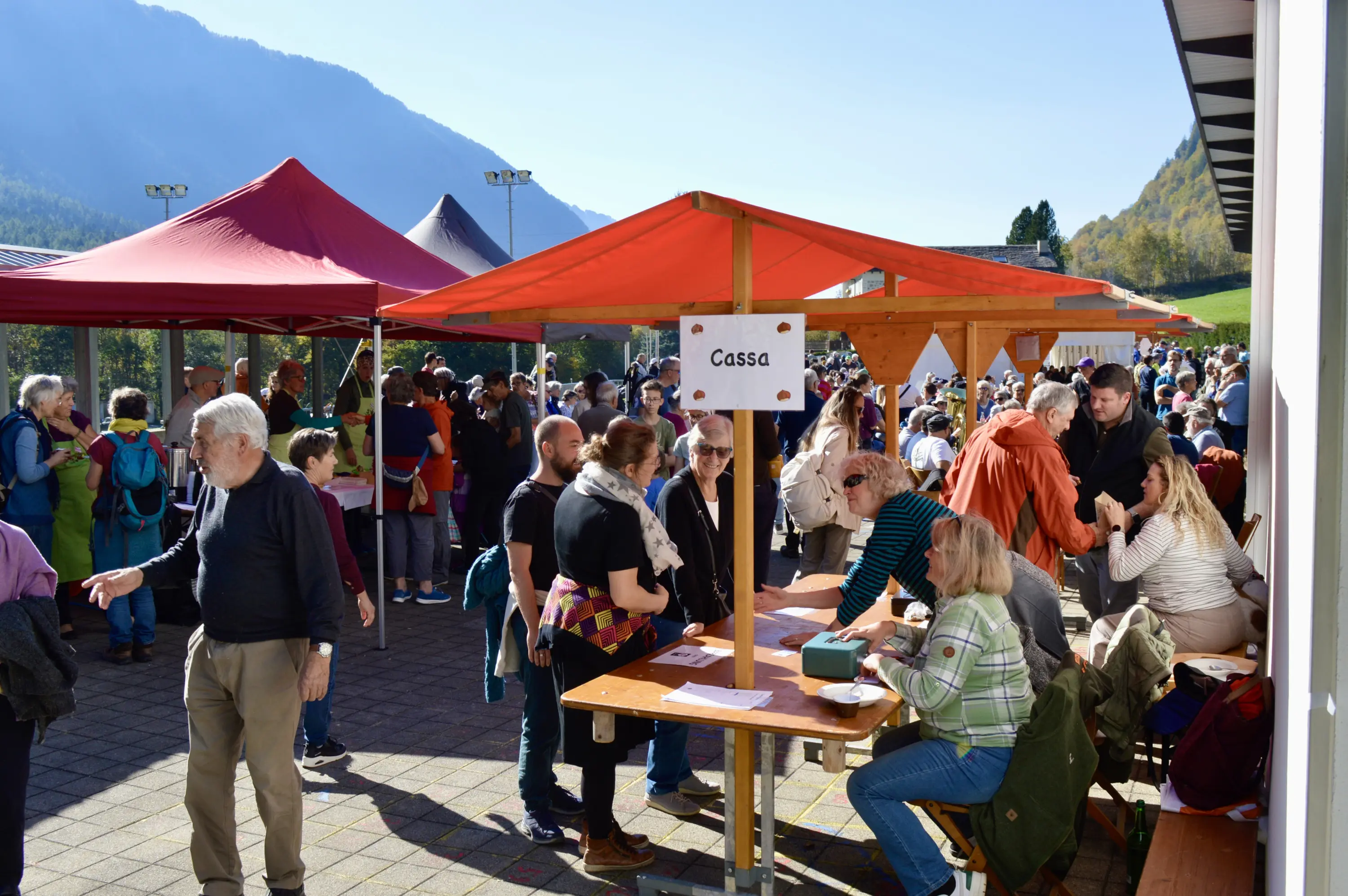 Una vivace scena di una fiera all'aperto con molte persone. Diverse bancarelle sono disposte, alcune con tende rosse. In primo piano si vede una cassa con un cartello che indica "Cassa". Sullo sfondo ci sono colline e alberi.