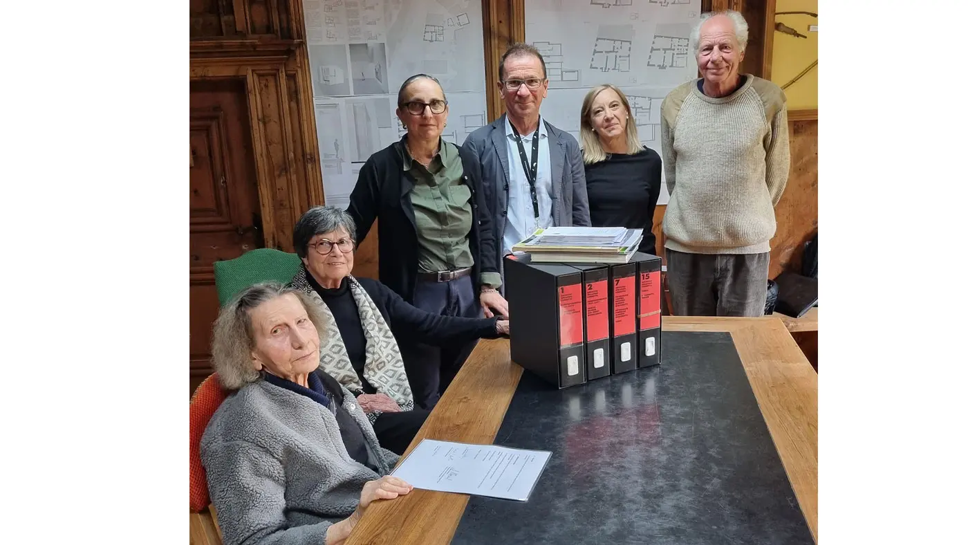 Un gruppo di sei persone si trova attorno a un tavolo. Alcuni di loro sorridono, mentre altri guardano verso la fotocamera. Sul tavolo ci sono alcuni documenti e tre raccoglitori con etichette rosse. Sullo sfondo, si vedono delle planimetrie appese al muro.