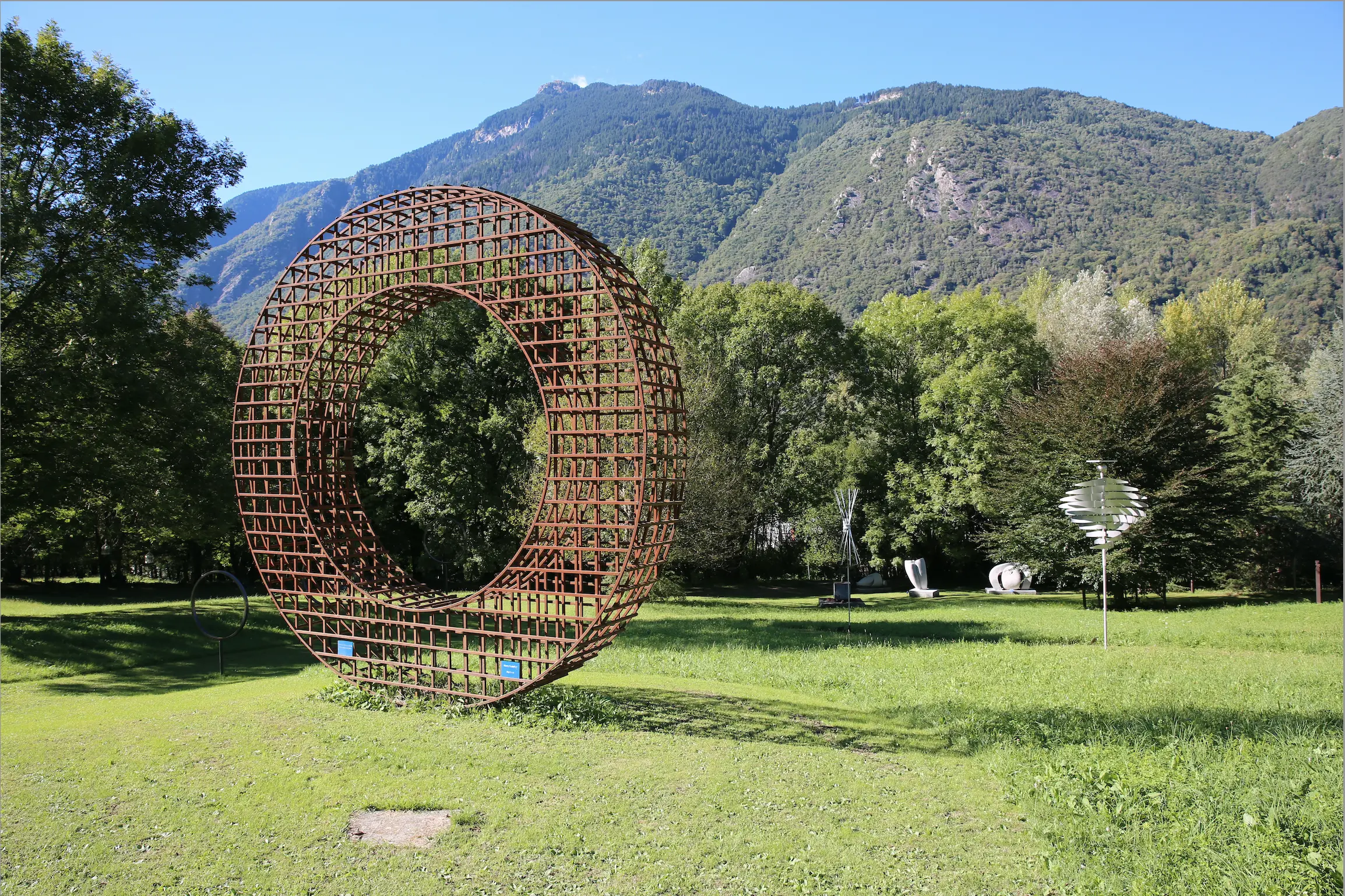 Scultura a forma di grande anello in ferro, immersa in un paesaggio verde con alberi e montagne sullo sfondo. Altre opere d'arte visibili in lontananza.