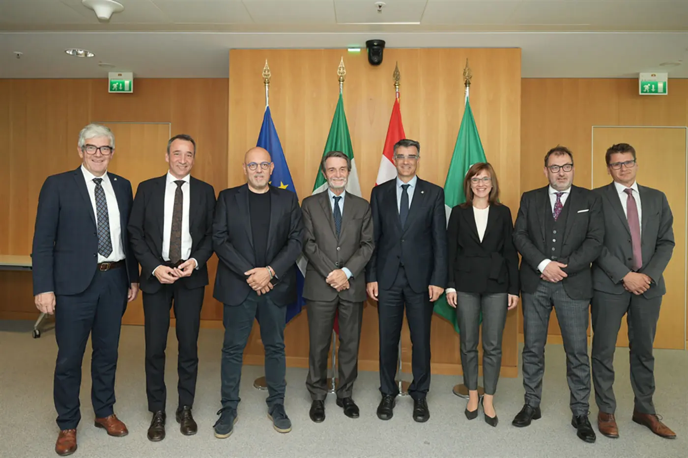 Un gruppo di sette persone in giacca e cravatta posa per una foto, sorridendo. Sullo sfondo sono visibili le bandiere dell'Unione Europea e di altri enti.