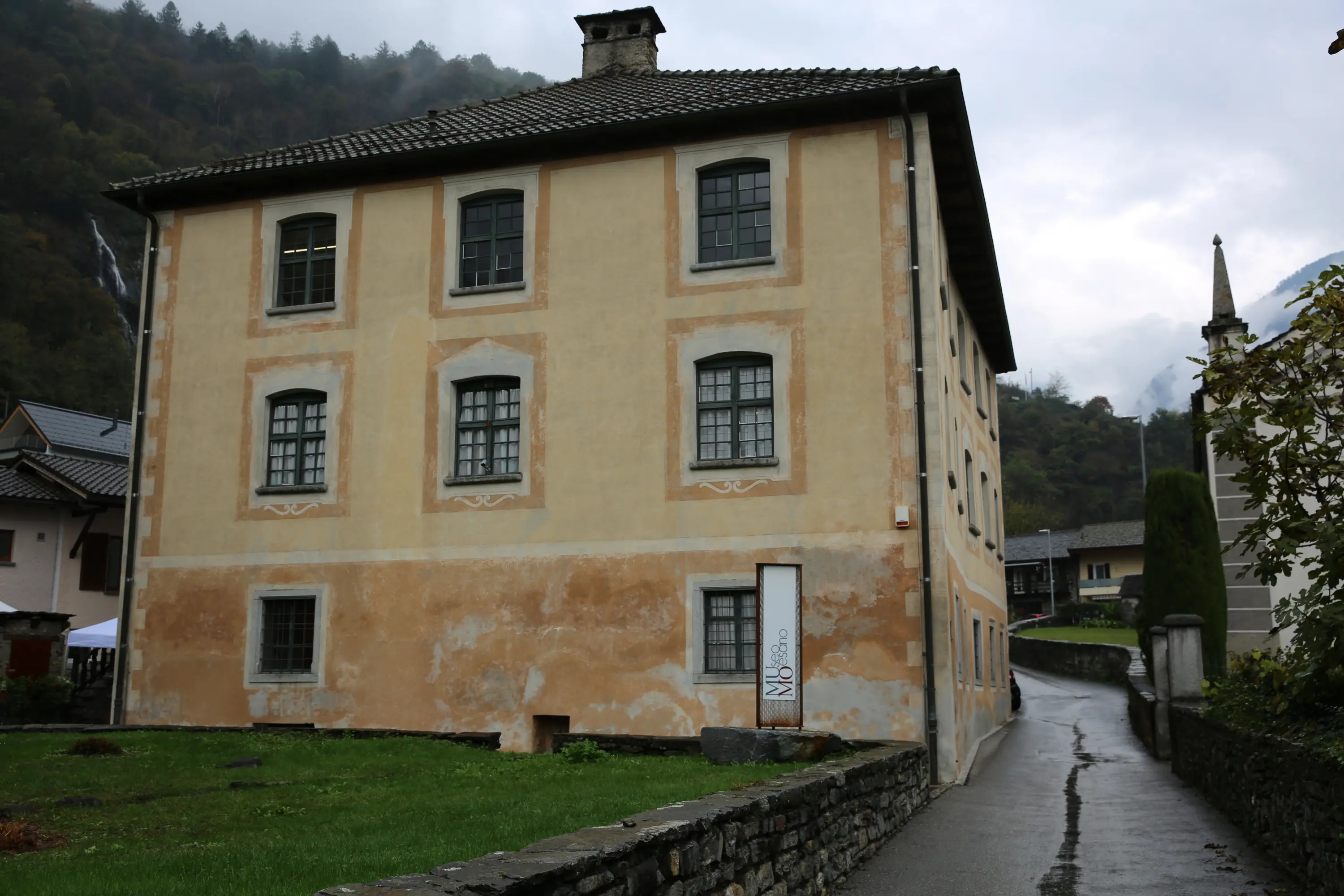 Edificio storico con pareti gialle e finestre con persiane, situato in una strada di montagna. Sul lato destro si intravede una chiesa e un paesaggio collinare.