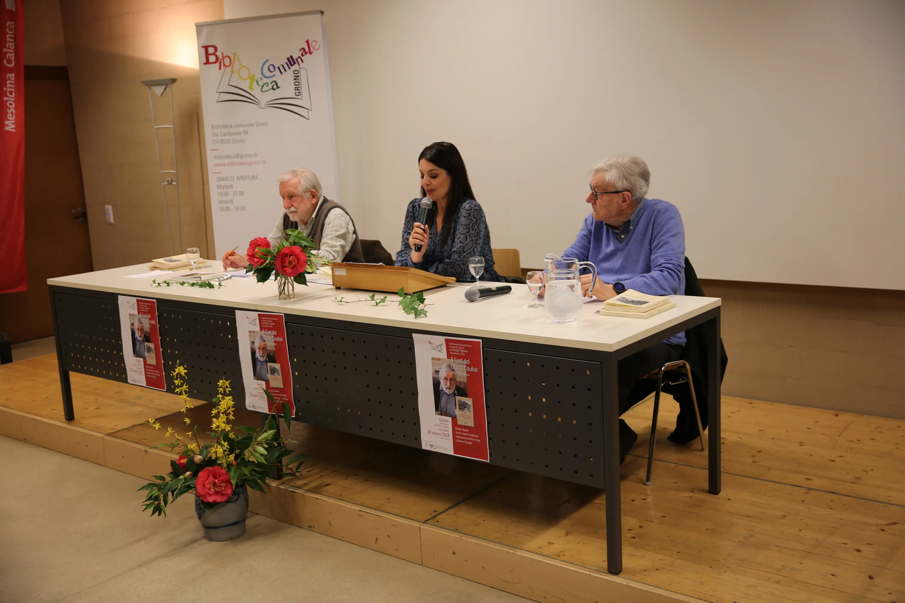 Tre relatori sono seduti a una tavola in un auditorium. Al centro, una donna legge da un testo, mentre gli altri due uomini la ascoltano. Sulle loro scrivanie ci sono libri e fiori freschi decorativi. Un poster è affisso sul muro dietro di loro.