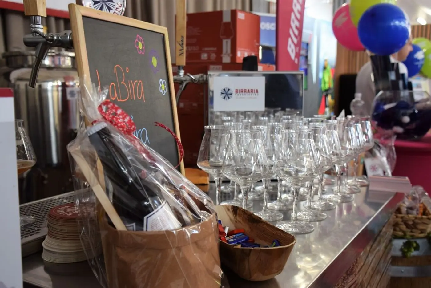 Una bancarella con una varietà di birre, bicchieri da degustazione trasparenti e una bottiglia in un cesto. Sullo sfondo si vedono decorazioni colorate e banner promozionali.