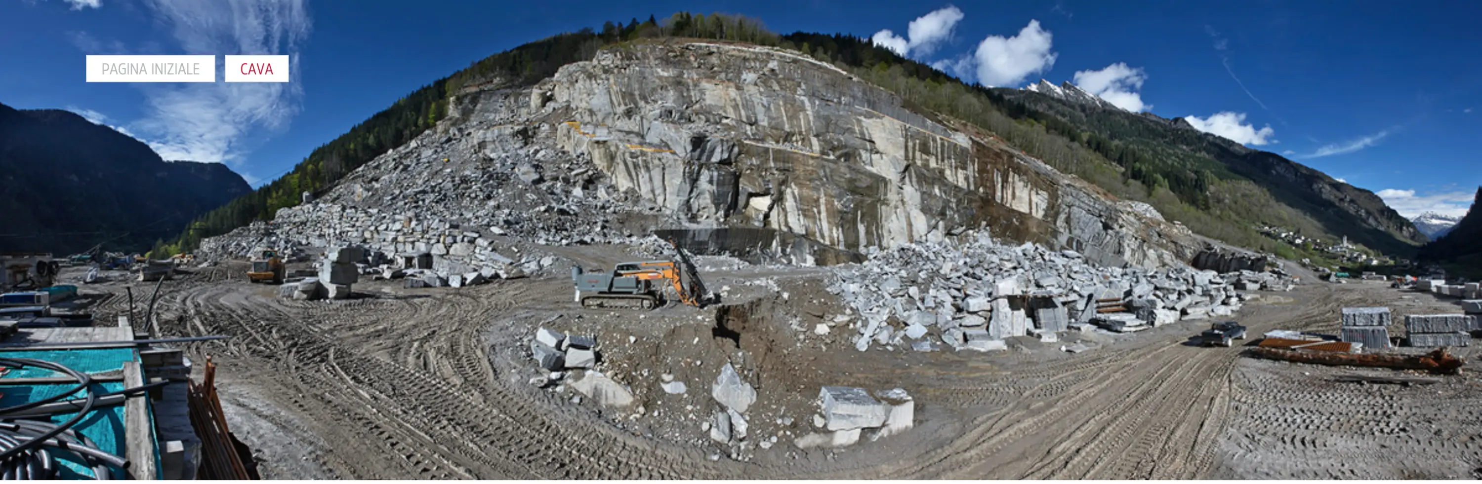 Lavori in una cava di pietra, con enormi blocchi di marmo ed attrezzature per l'estrazione. Sullo sfondo, una montagna e un cielo blu con nuvole.
