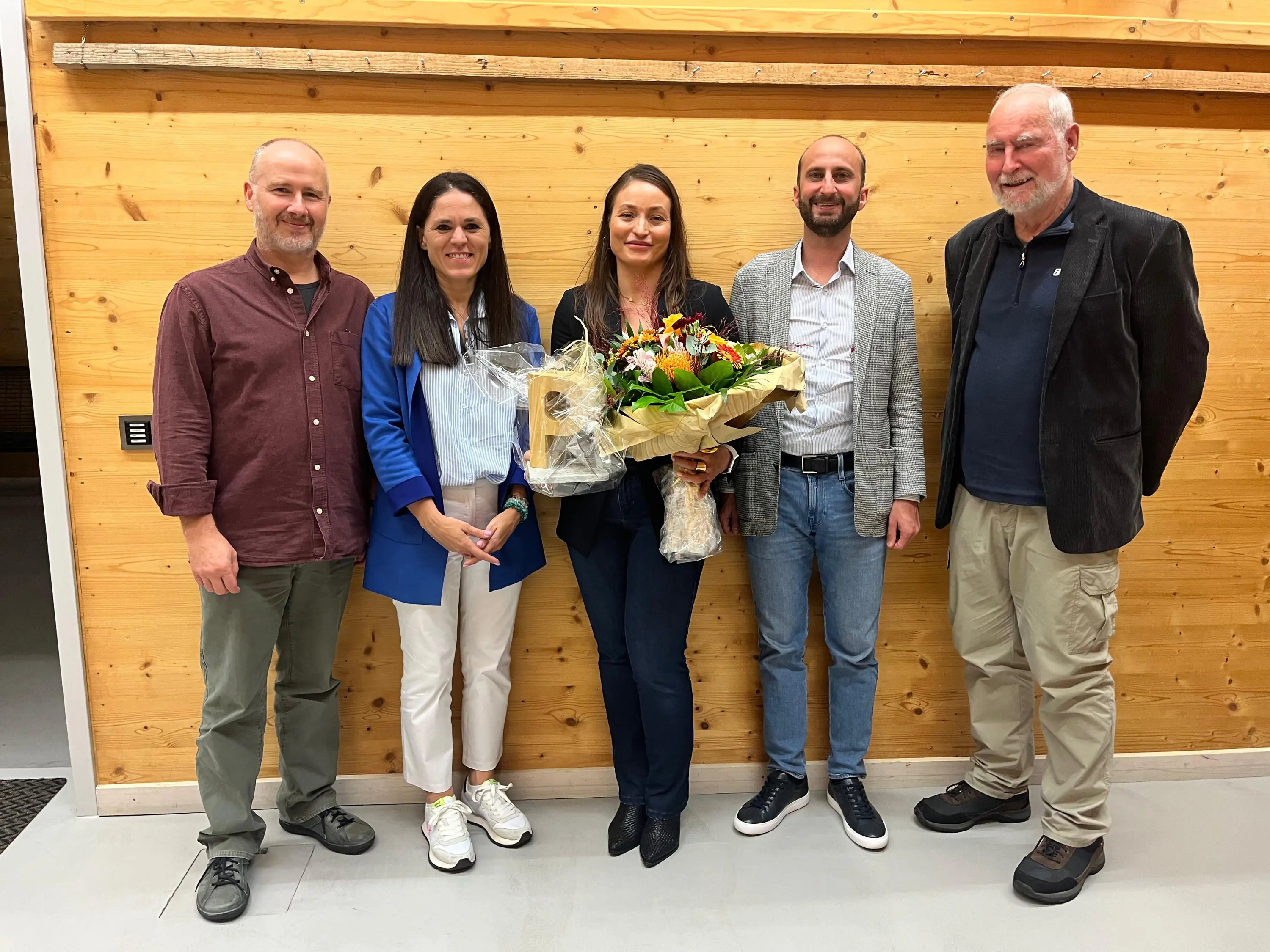 Quattro persone in piedi insieme, sorridenti, con una donna al centro che tiene un grande mazzo di fiori. Lo sfondo è di legno chiaro. Gli altri indossano abbigliamento informale e formale.