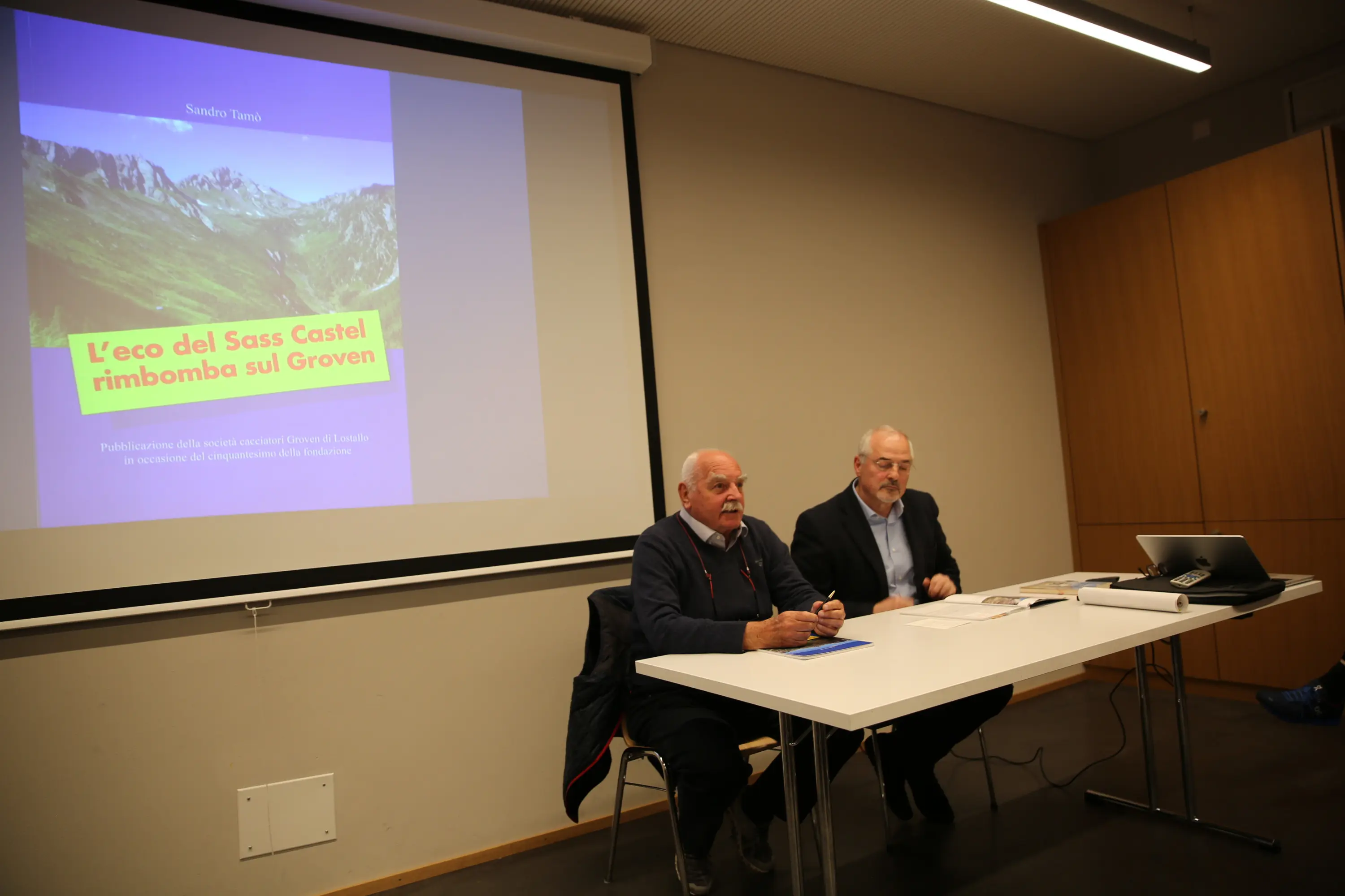 Due relatori discutono durante un evento di presentazione. Sullo sfondo si vede un poster con il titolo "L'eco del Sass Castel rimbomba sui Groven". Sono presenti un tavolo con computer e documenti.