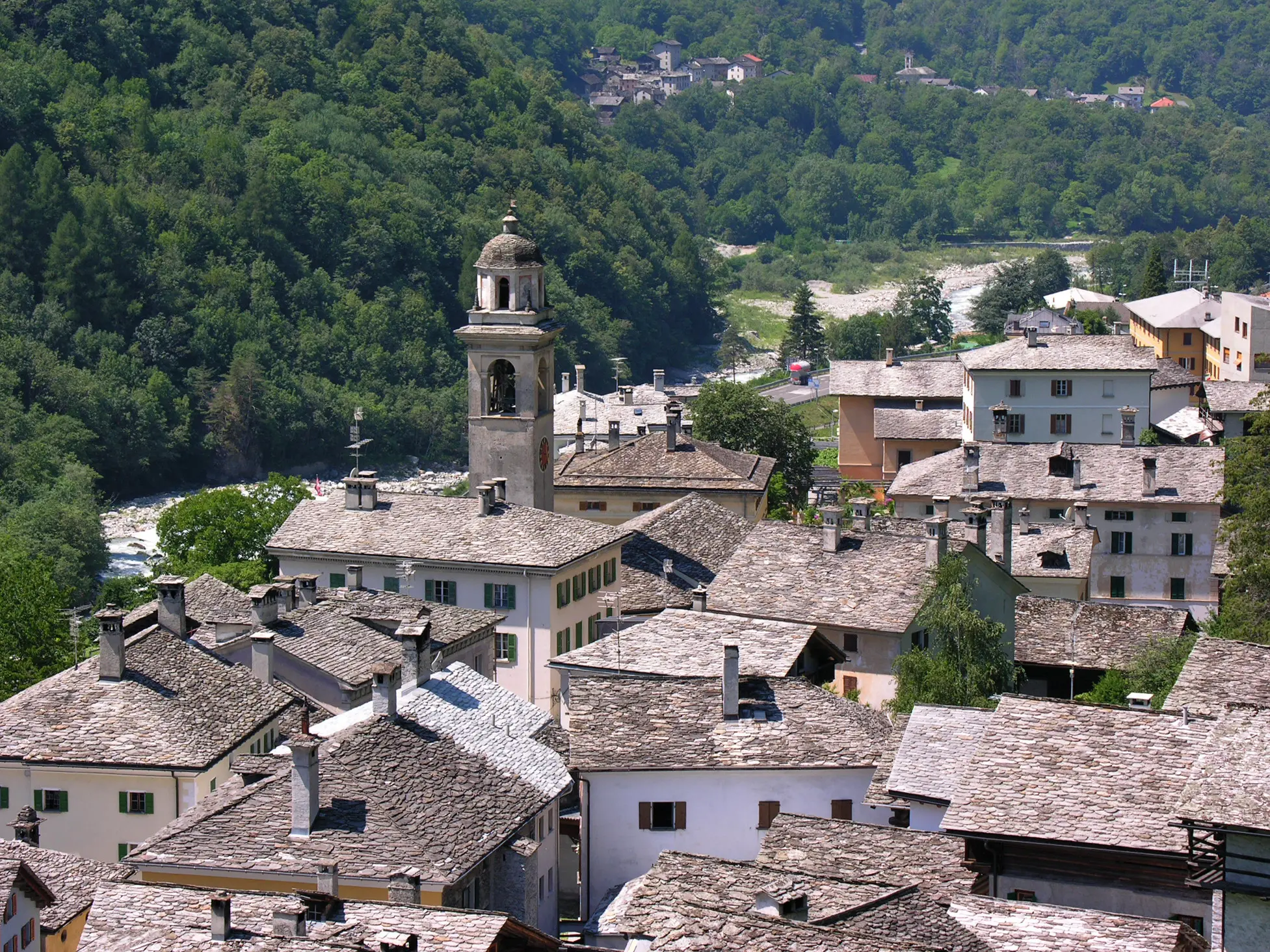 Vista panoramica di un villaggio montano con tetti in pietra e una torre campanaria. Circondato da alberi verdi e colline, il fiume scorre in lontananza.