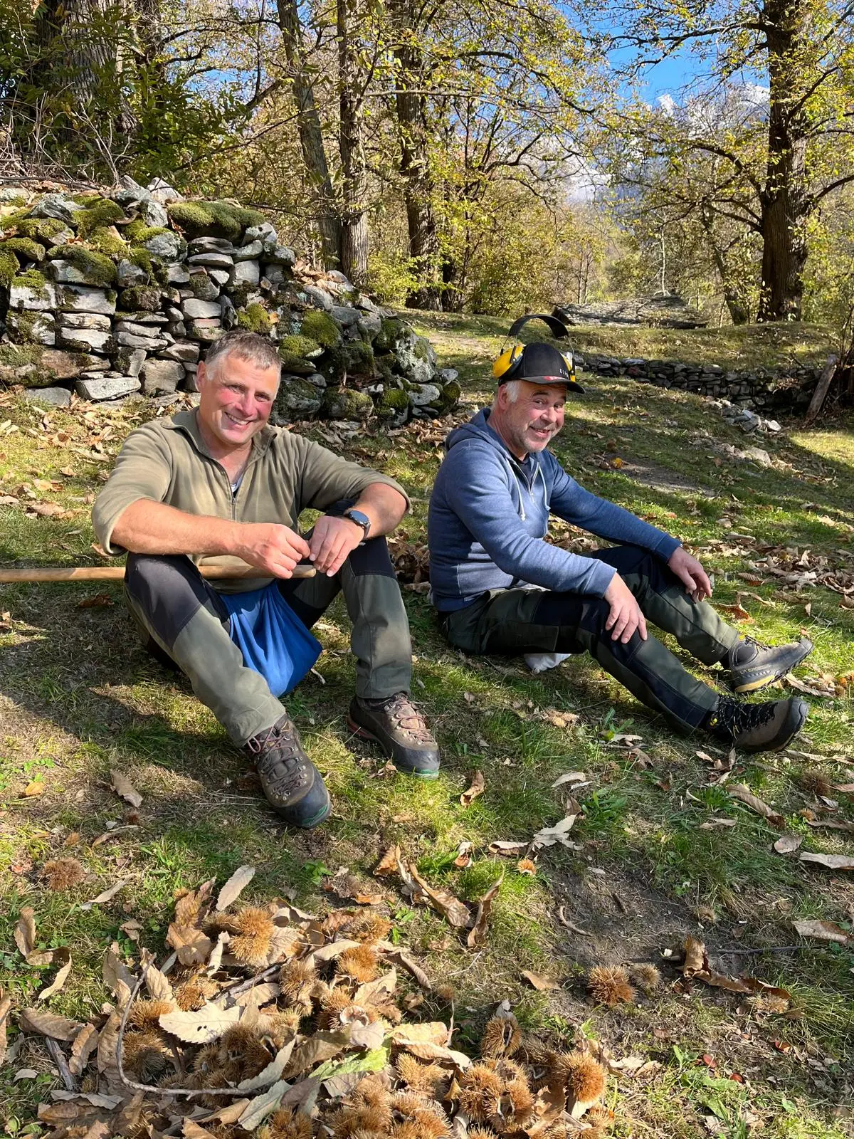 Due uomini sorridenti seduti sull'erba in un bosco, circondati da foglie e castagne. Sullo sfondo, un muro di pietra e alberi con foglie autunnali.