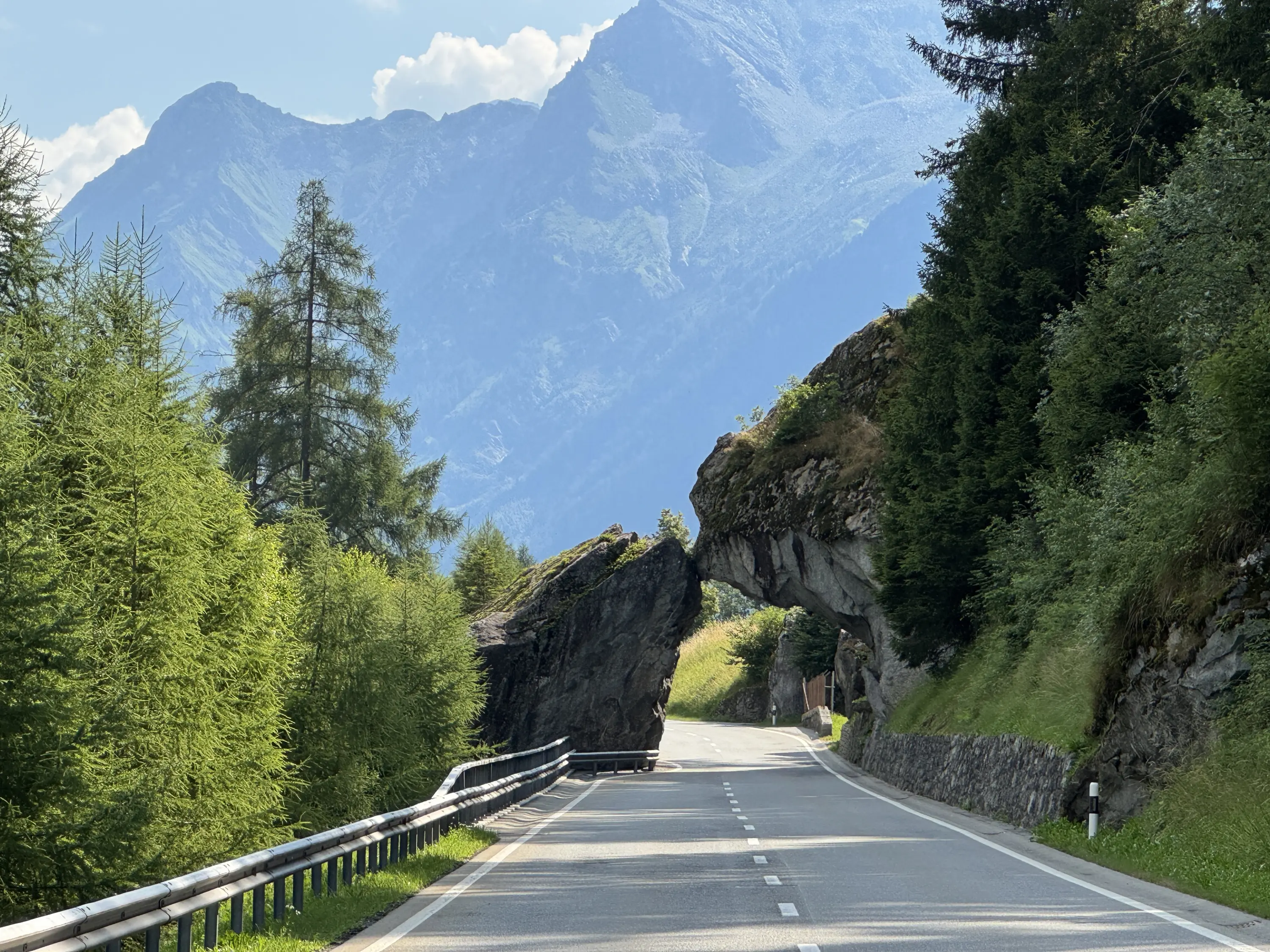 Una strada sinuosa attraversa un'apertura rocciosa, circondata da alberi verdi e montagne maestose sullo sfondo.