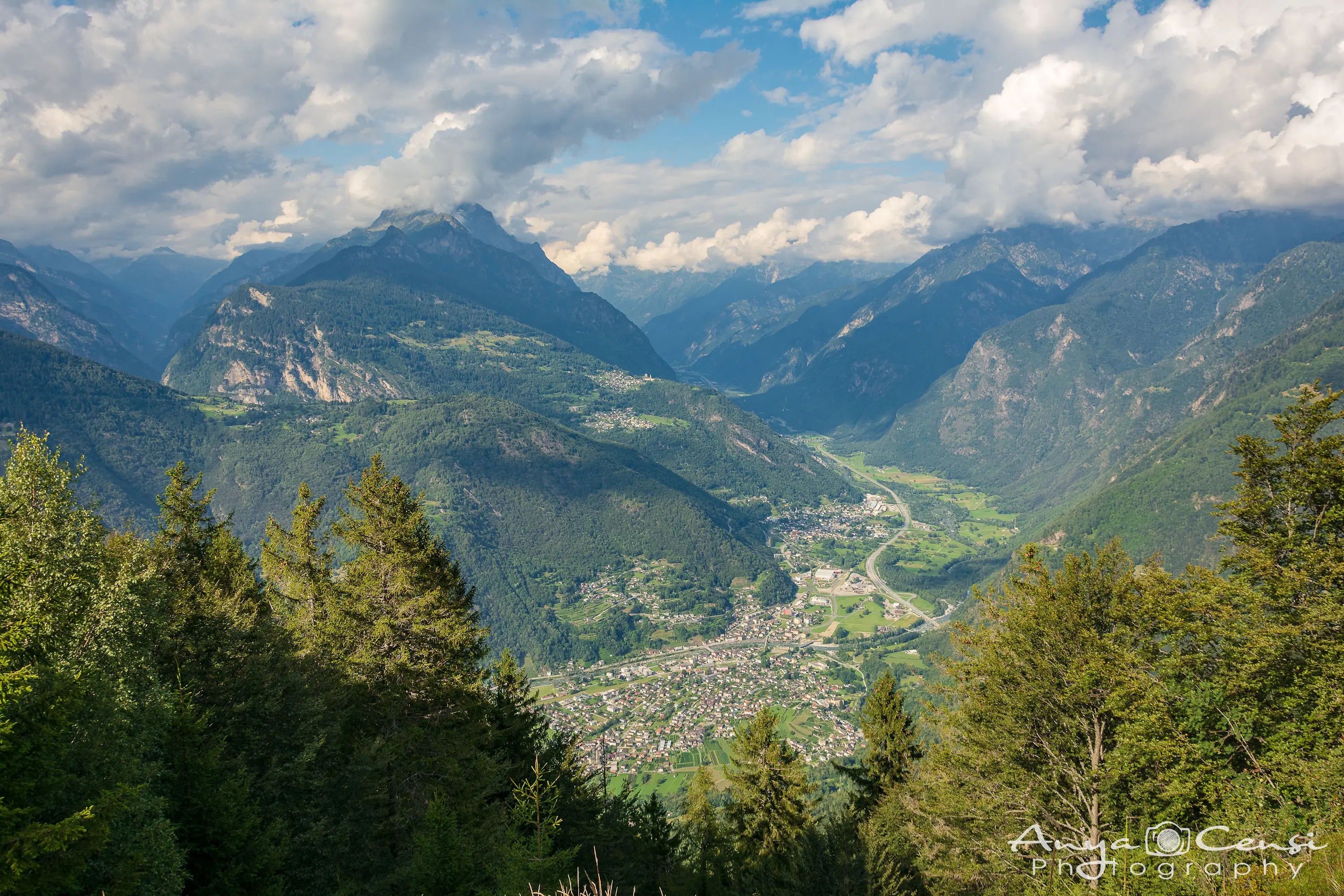 Valle verdeggiante circondata da montagne maestose. Nuvole sparse nel cielo azzurro. Un paese si trova in basso, circondato da alberi.