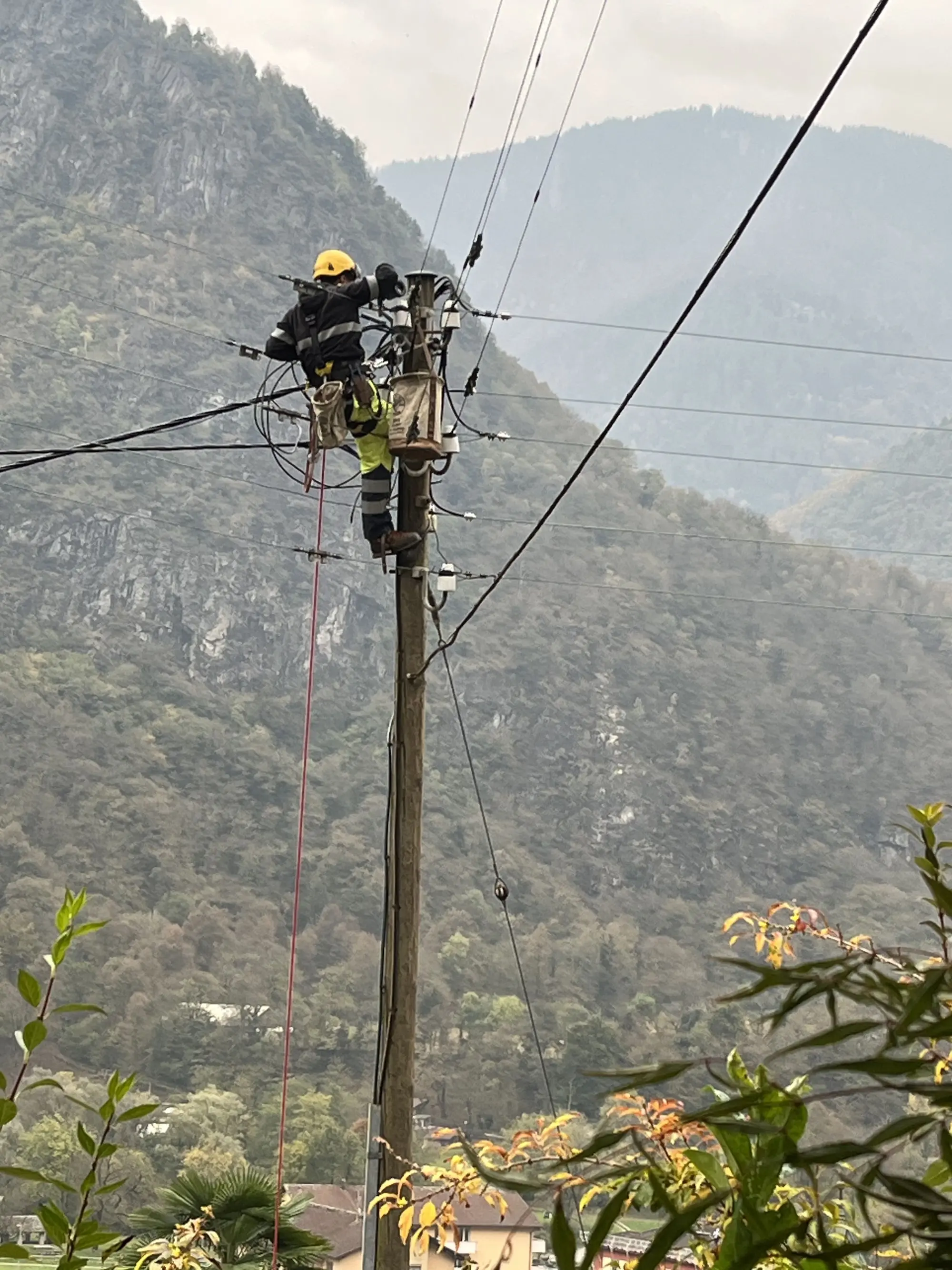 Un tecnico in sicurezza, con elmetto giallo, lavora su un palo della luce, mentre sistema i cavi elettrici in un'area montuosa.
