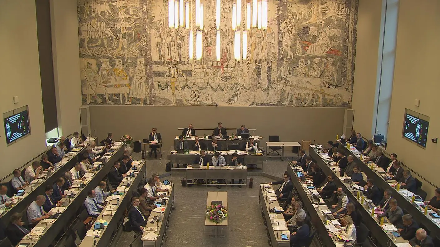 Una sala conferenze con un gruppo di persone sedute attorno a tavoli disposti a forma di U. Un grande arazzo decorativo copre la parete sullo sfondo, mentre schermi e fiori adornano i tavoli.