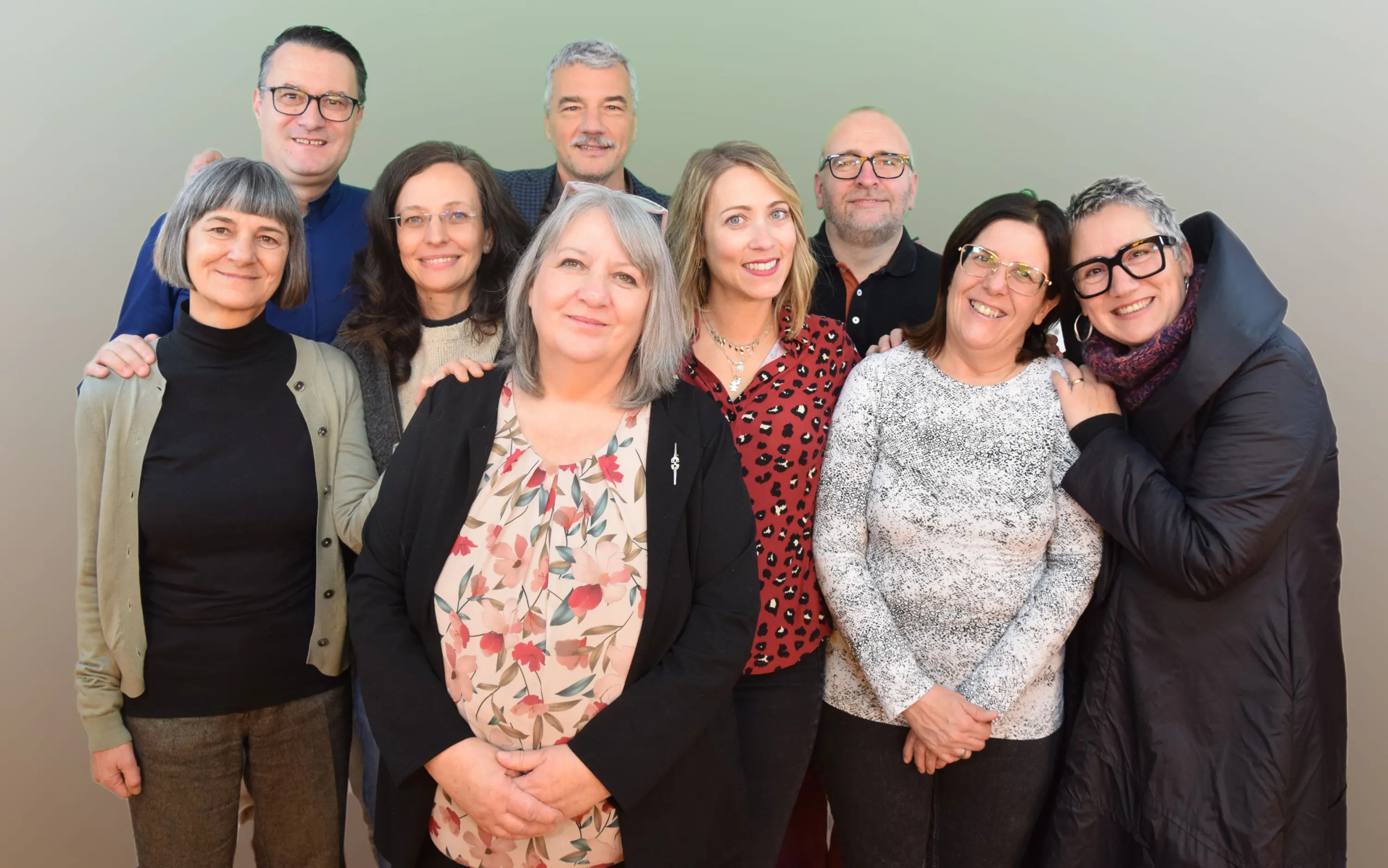 Gruppo di persone sorridenti in un ambiente ben illuminato, alcune con occhiali. Abbigliamento vario, in un mix di stili e colori. Sfondo sfumato chiaro.