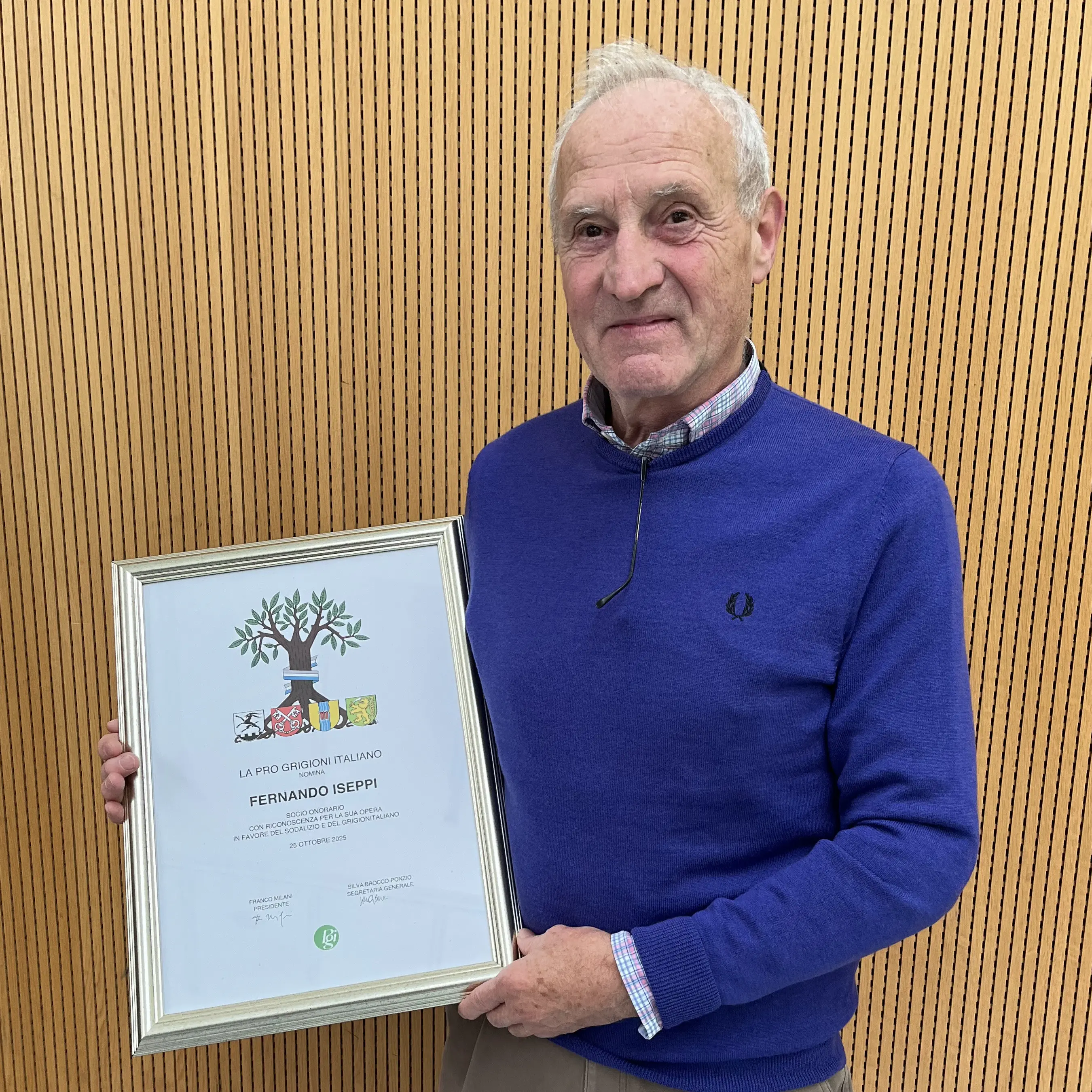 Uomo anziano con cappotto blu tiene un certificato incorniciato. Sul certificato c'è un albero stilizzato e il nome "Fernando Iseppi". Sullo sfondo, un muro di legno.