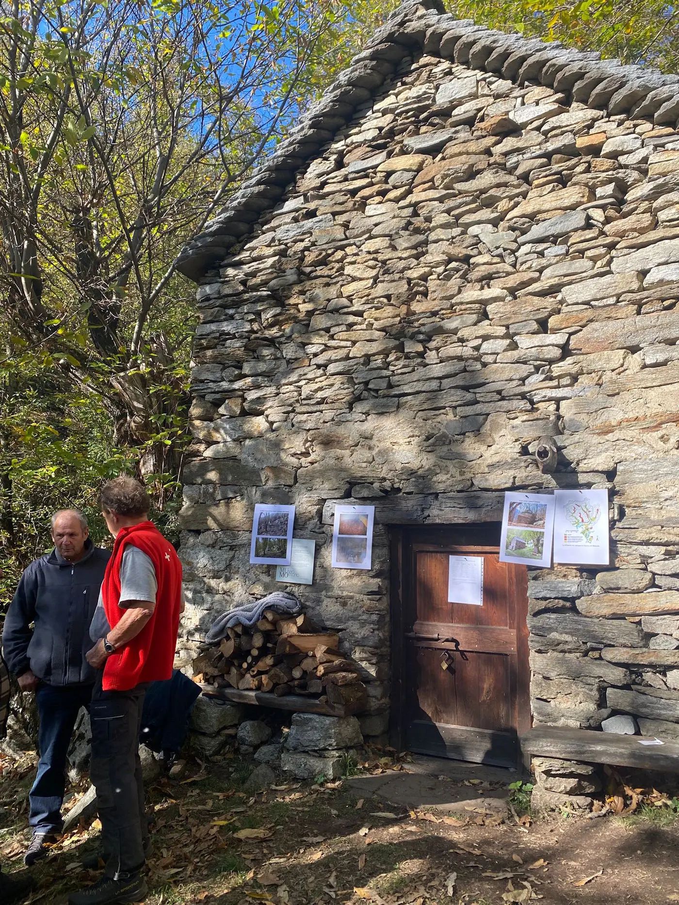 Due uomini si trovano di fronte a una casa in pietra con un tetto di sassi. La parete è adornata da fotografie appese. Accanto alla porta di legno, c'è una catasta di legna. Gli alberi e il cielo blu si intravedono sullo sfondo.