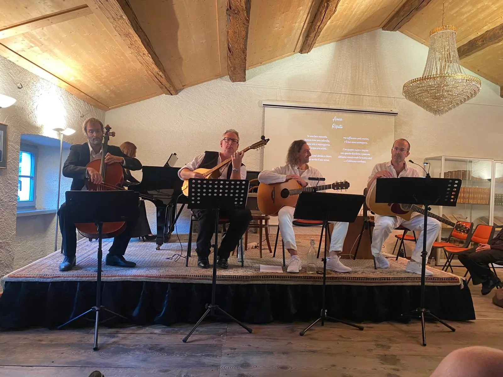 Quattro musicisti su un palco, ognuno con il proprio strumento: un violoncellista, un chitarrista e due musicisti con chitarre acustiche, mentre si esibiscono in un ambiente intimo con travi a vista e un lampadario.