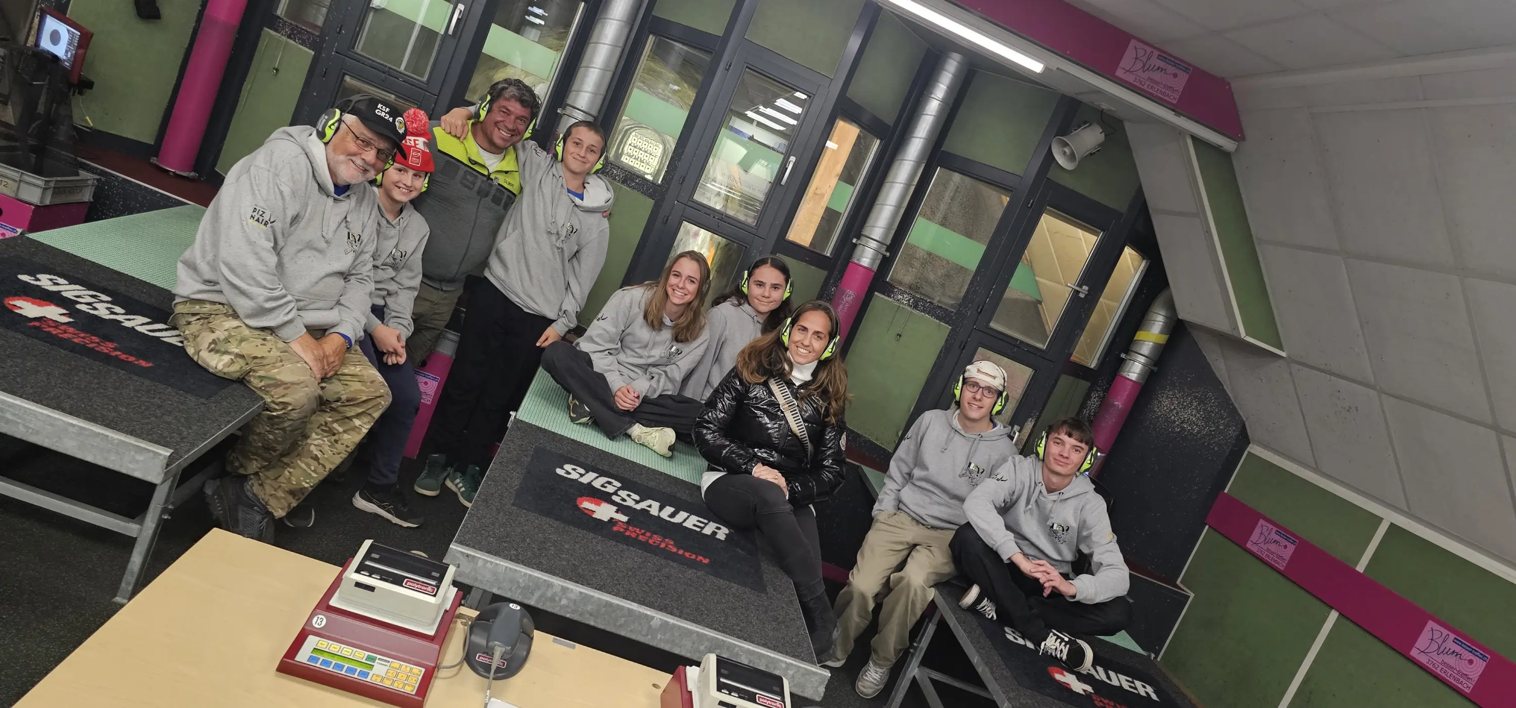 Gruppo di otto persone sorridenti, sedute su tavoli in una struttura di addestramento per il tiro. Ognuno indossa un maglione grigio, mentre alcune persone portano cappellini. Il contesto è moderno, con pareti colorate e attrezzature per il tiro visibili sullo sfondo.