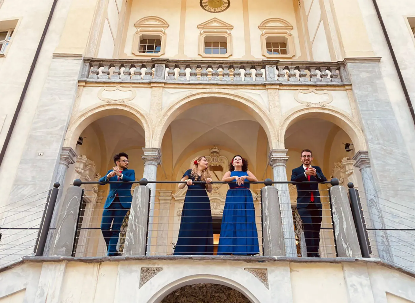 Quattro persone sorridenti in abiti eleganti, due donne e due uomini, posano su un balcone con archi decorativi. Il loro sfondo è un palazzo storico con dettagli architettonici raffinati.