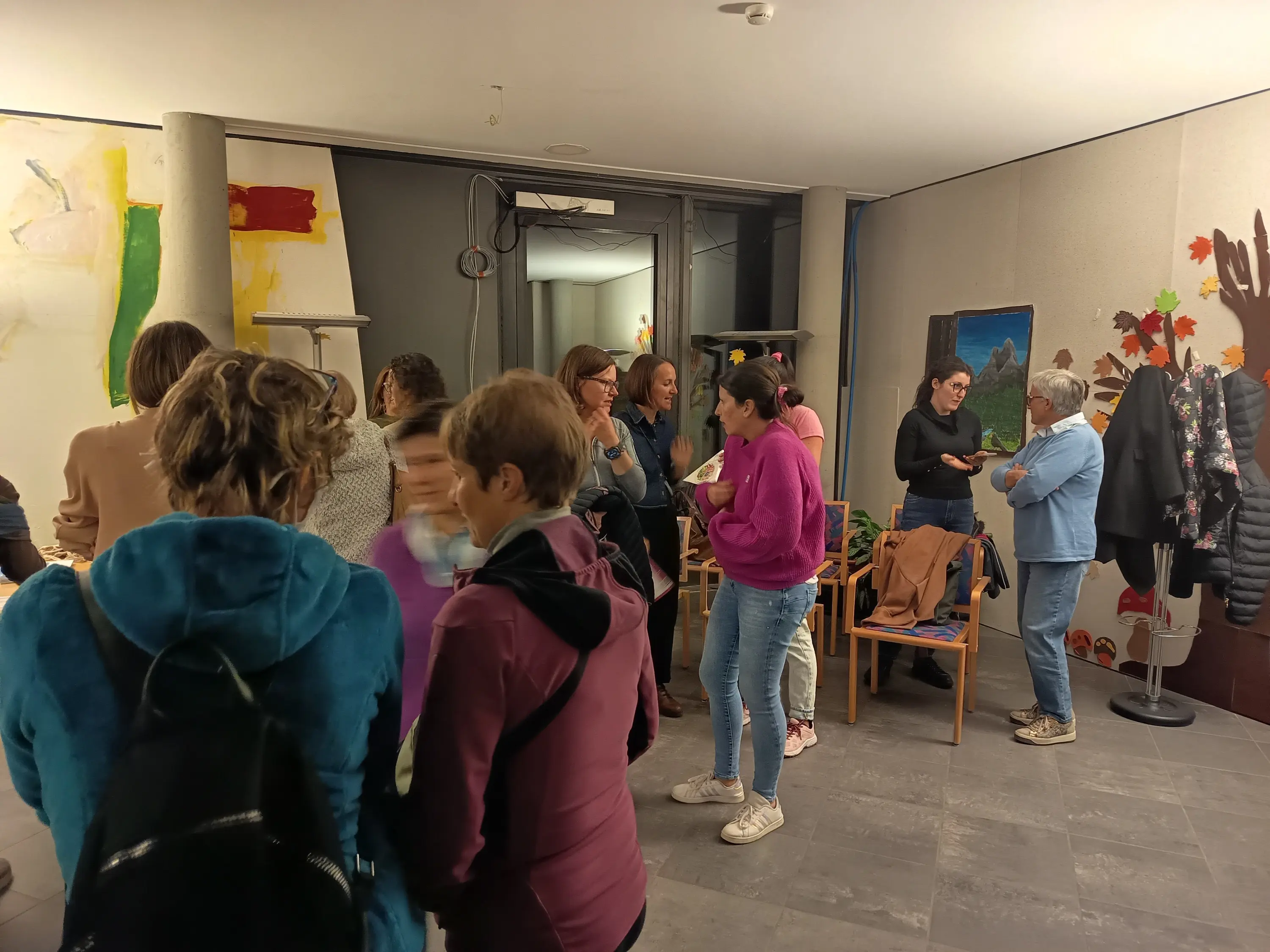 Un gruppo di persone si riunisce in un ambiente interno, con opere d'arte alle pareti e alcune sedie. Alcuni scambiano idee, mentre altri sono assorti nei loro telefoni.
