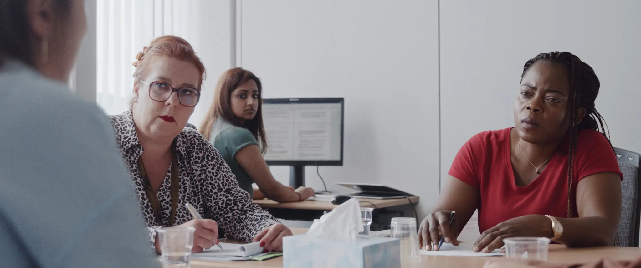 Un gruppo di donne sedute attorno a un tavolo in una riunione. Una donna distinta con occhiali fissa la telecamera con un'espressione seria, mentre accanto a lei un'altra donna con un top rosso ascolta attentamente. In background, un'altra donna è seduta davanti a un computer. Su un tavolo ci sono fogli, bicch