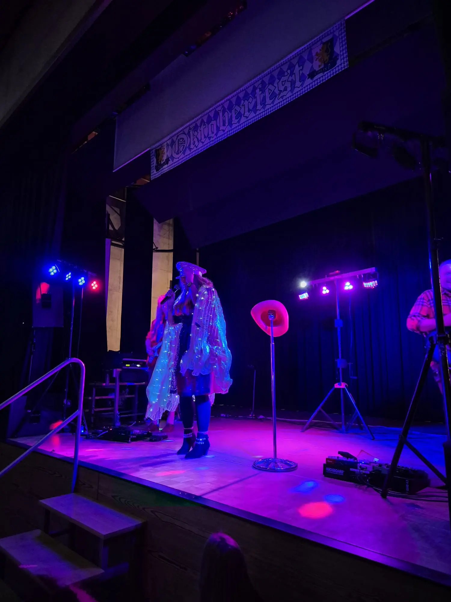 Un performer sul palco indossa un costume colorato e trasparente, illuminato da luci blu e viola. Sullo sfondo, si vede un arco con la scritta "Oktoberfest". Strumenti musicali sono presenti sul palco.
