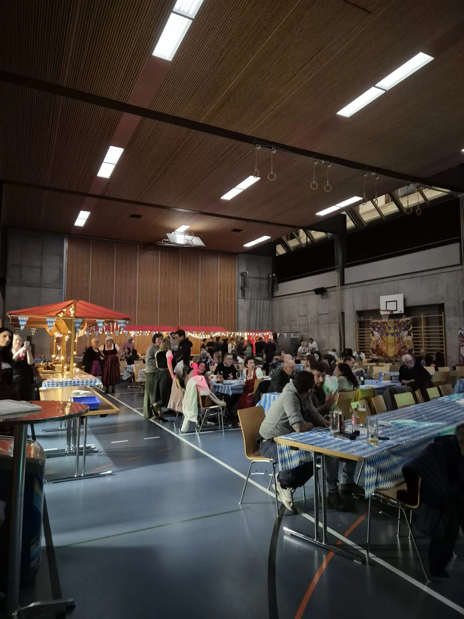 Una sala gremita di persone durante una festa, con tavoli decorati e bandierine colorate. Alcuni partecipanti indossano abiti tradizionali e c'è un'area con un bancone in legno.