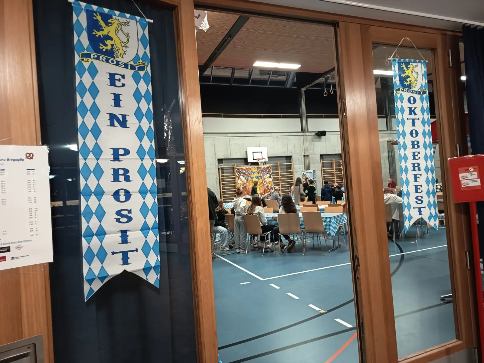Insegne che annunciano la festa dell'Oktoberfest, appese in una sala con tavoli e partecipanti. Si intravede un palcoscenico con decorazioni festose, mentre il locale è allestito in stile bavarese.