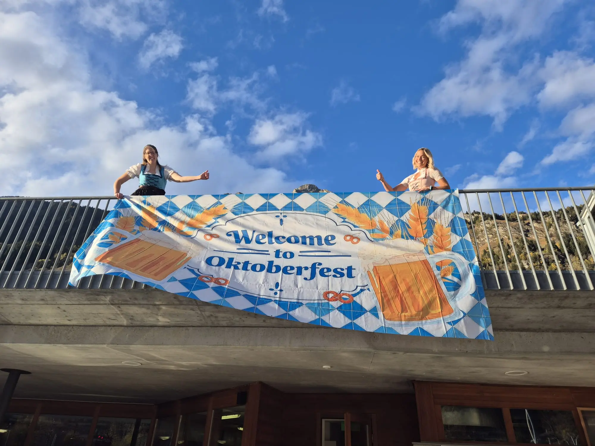 Due donne sorridenti posano con un grande striscione che dice "Welcome to Oktoberfest", decorate con immagini di boccali di birra e spighe di grano. Il cielo azzurro e nuvoloso fa da sfondo.
