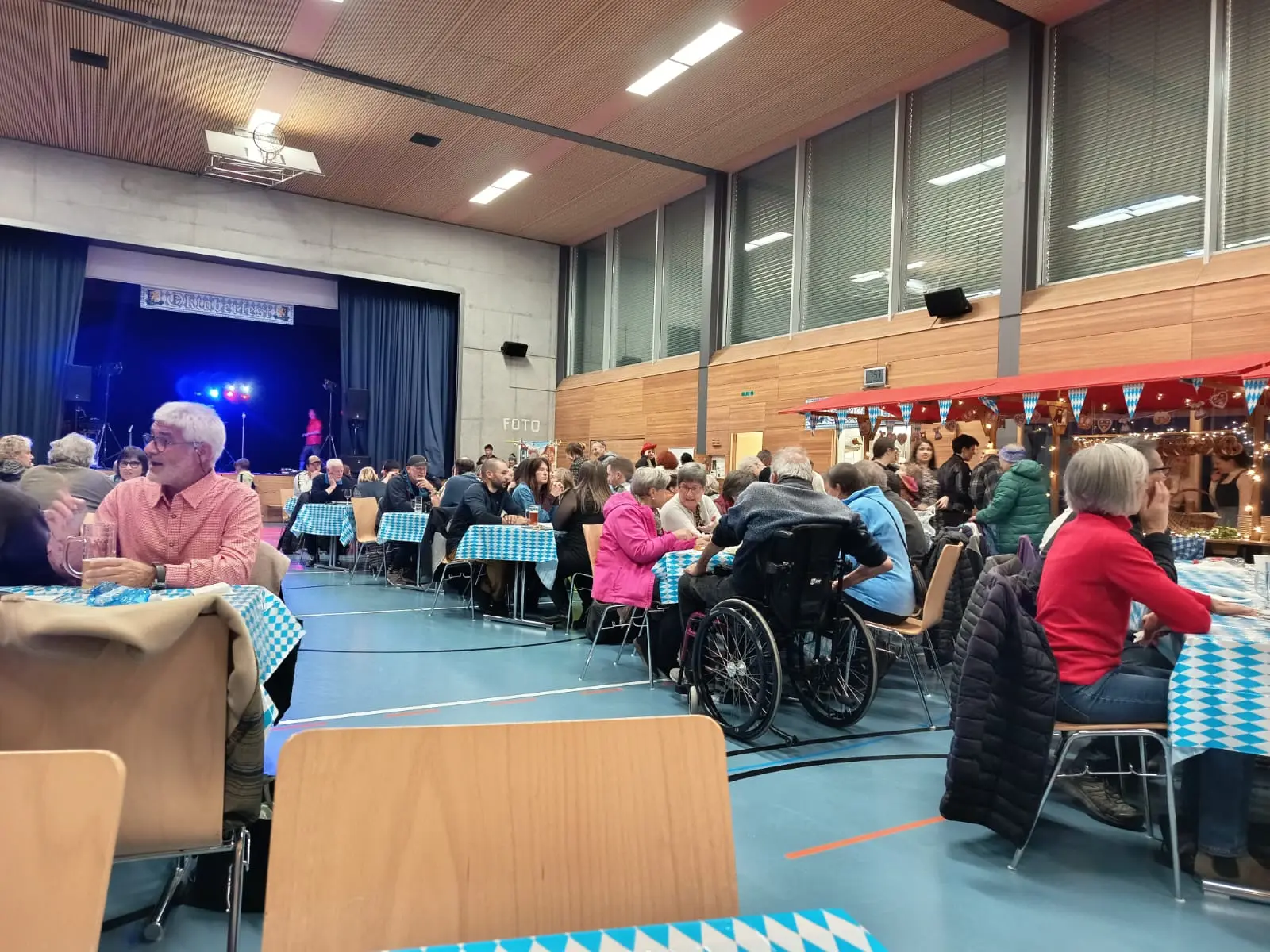 Una sala affollata con persone sedute a tavoli decorati, persone che conversano e un palco sullo sfondo illuminato.