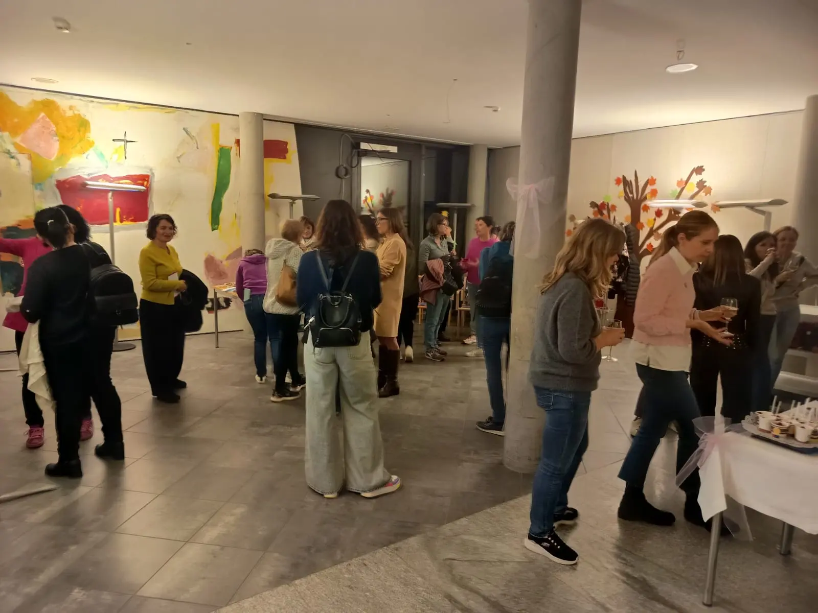 Un gruppo di persone si riunisce in un ambiente artistico e moderno, circondato da opere d'arte colorate. Alcuni partecipanti conversano, altri gustano bevande e stuzzichini, mentre l'atmosfera è vivace e sociale.