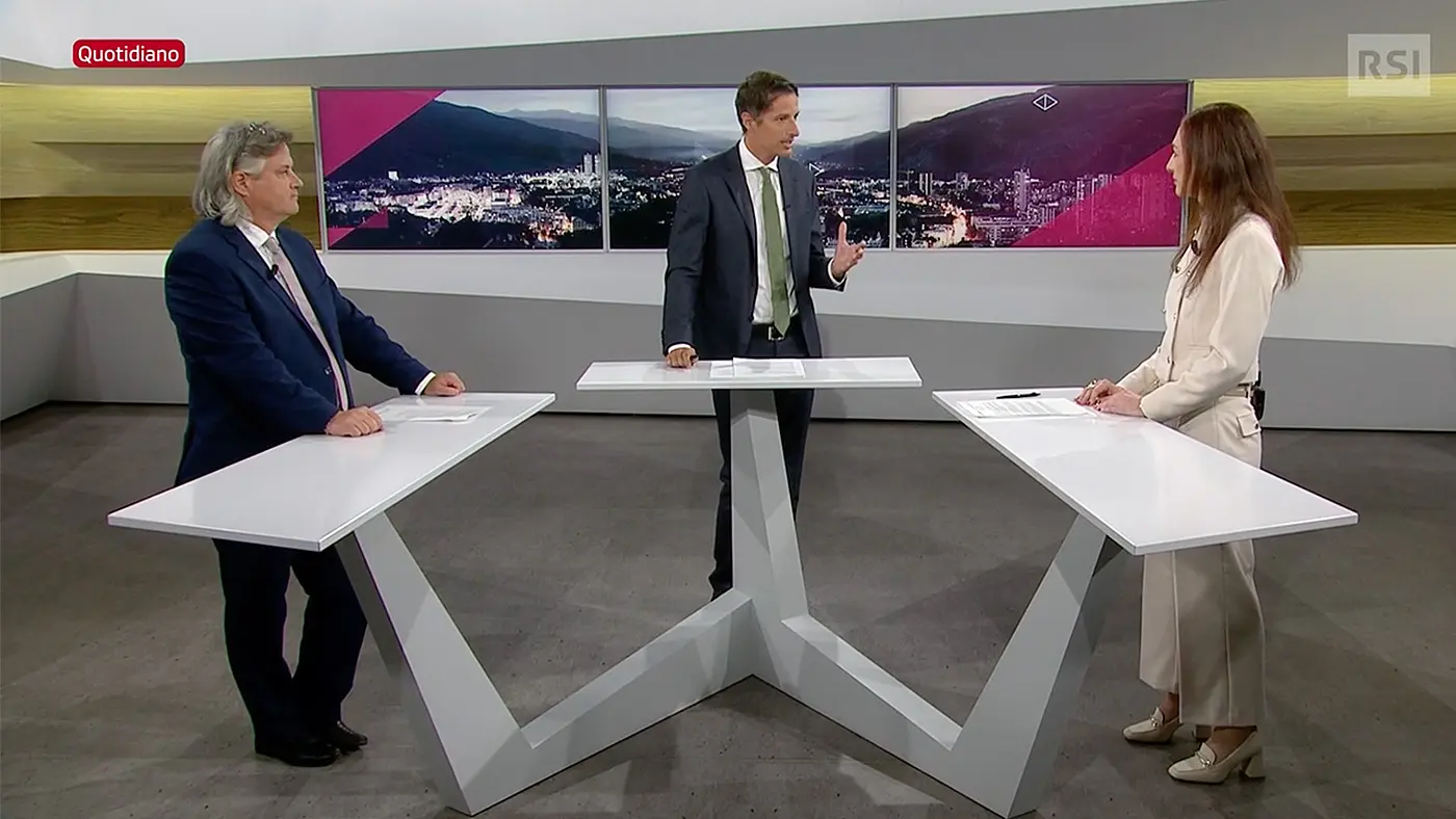 Tre persone in un set televisivo discutono. Due presentatori, uno a sinistra con capelli lunghi e grigi, l'altro a destra con un abito chiaro, ascoltano un moderatore al centro. Sullo sfondo si vedono immagini di una città.