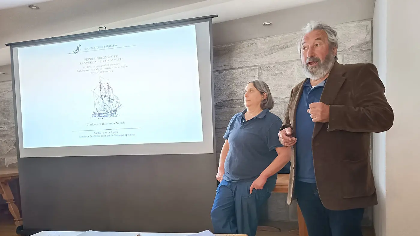 Due persone in un ambiente interno. Un uomo con giacca scura e capelli brizzolati parla di fronte a un proiettore, mentre una donna con capelli lunghi e grigi ascolta. Sullo sfondo, una presentazione è visibile su uno schermo.