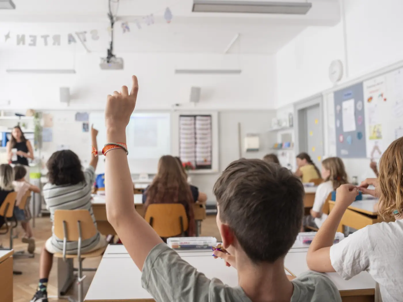 Una classe di studenti alzano le mani, seduti ai banchi. In primo piano, un ragazzo con la mano alzata. La docente spiega davanti a una lavagna. Diversi materiali didattici sono sparsi sui banchi.