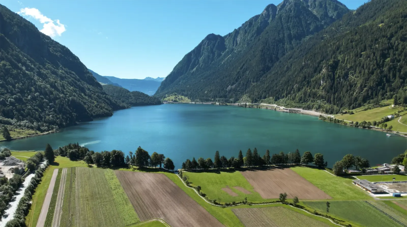 Una vista panoramica di un lago circondato da montagne verdi e ripide, con campi coltivati nel primo piano. Il cielo è sereno e blu.