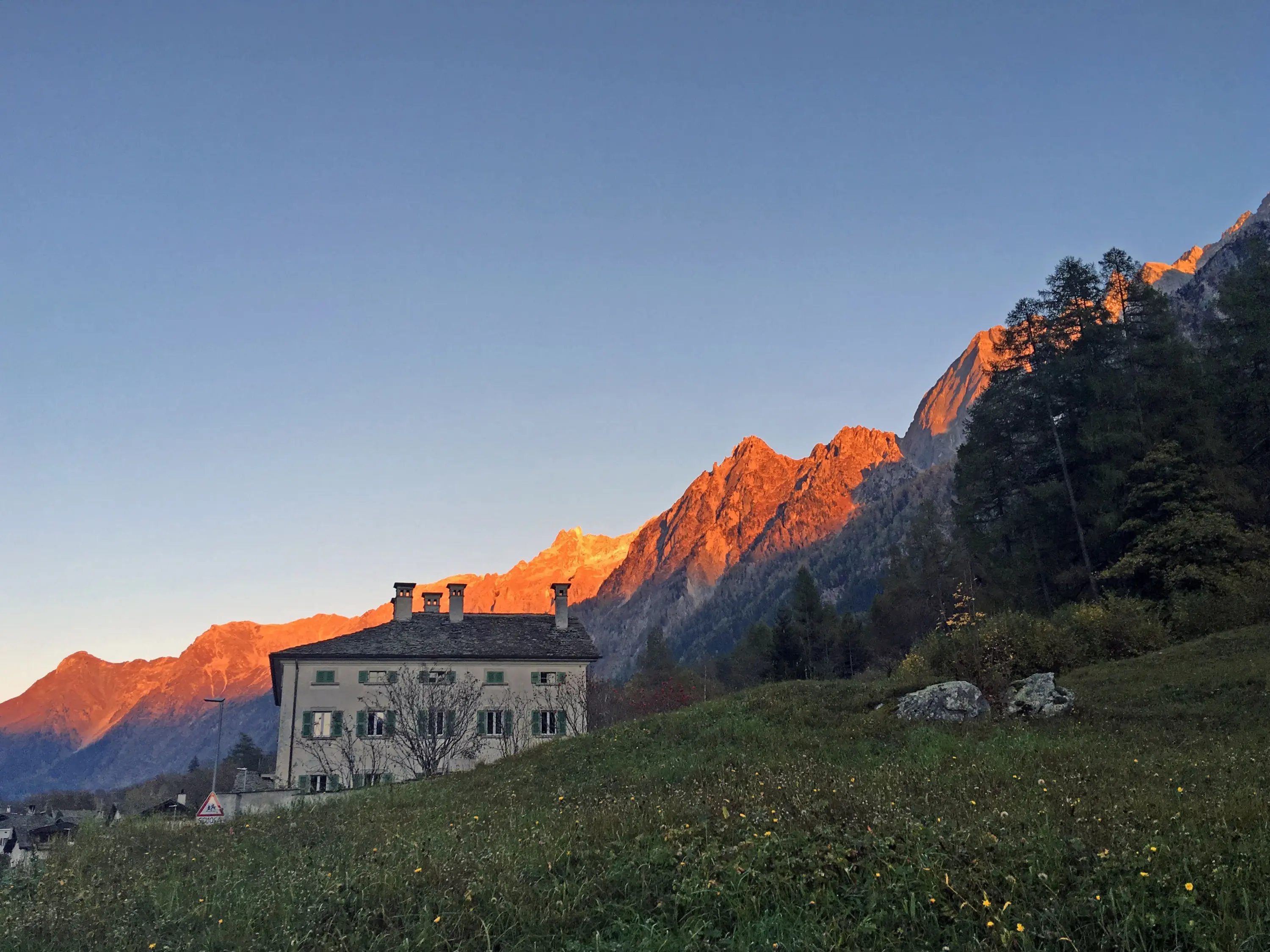 Una casa storica con pareti bianche si trova in primo piano, circondata da prati verdi e montagne imponenti illuminate dalla luce dorata del tramonto. Il cielo è sereno e di un blu profondo.