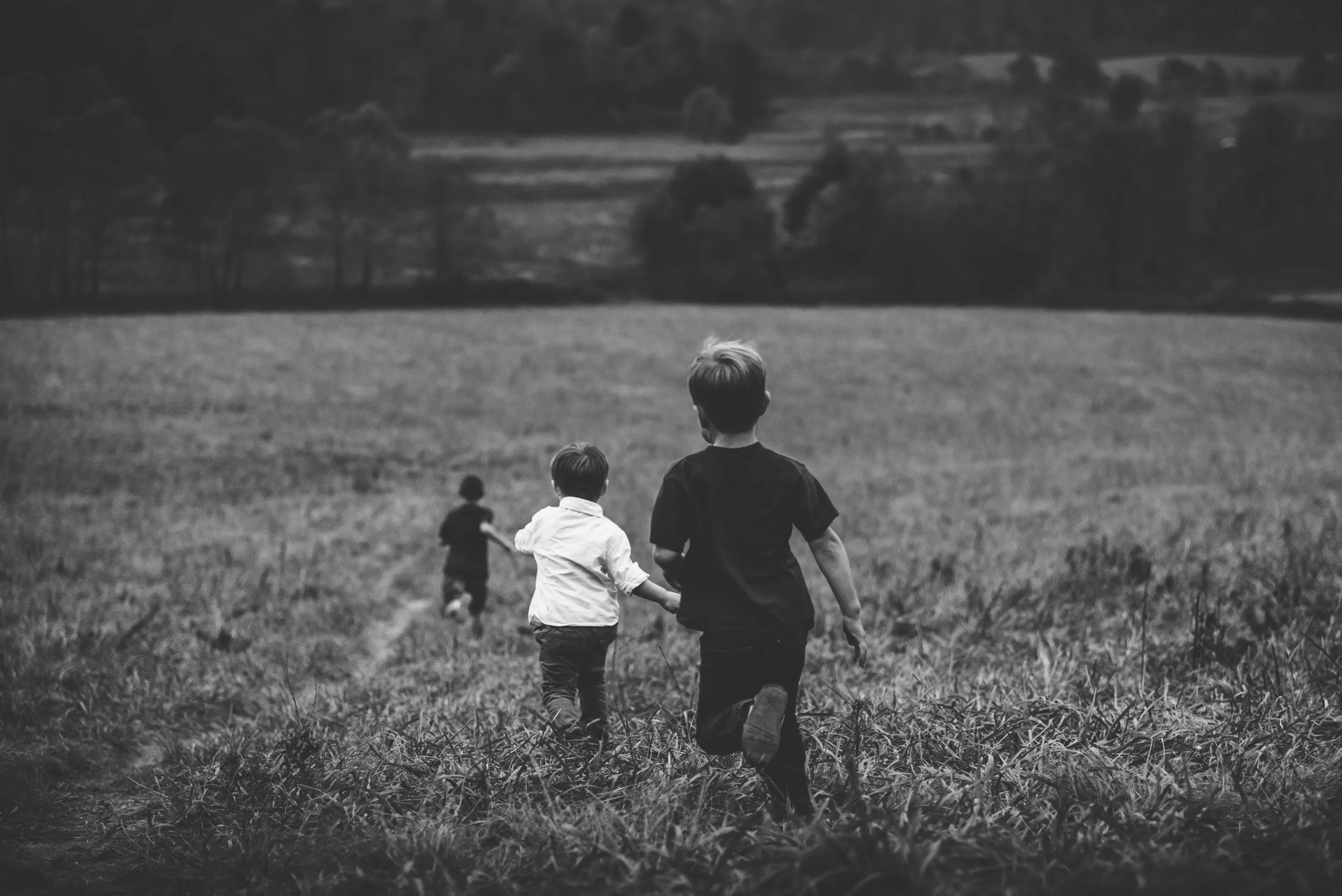 Due bambini corrono attraverso un campo verde, mentre un terzo bambino è leggermente più indietro. Il paesaggio è collinare e la scena è in bianco e nero.