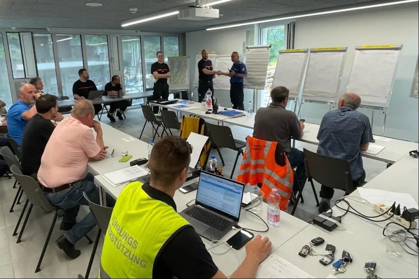 Un gruppo di uomini partecipa a un incontro formativo in una sala moderna. Alcuni sono seduti a un tavolo, mentre altri stanno presentando. Sono presenti lavagne bianche e dispositivi elettronici. L'atmosfera è di concentrazione e collaborazione.