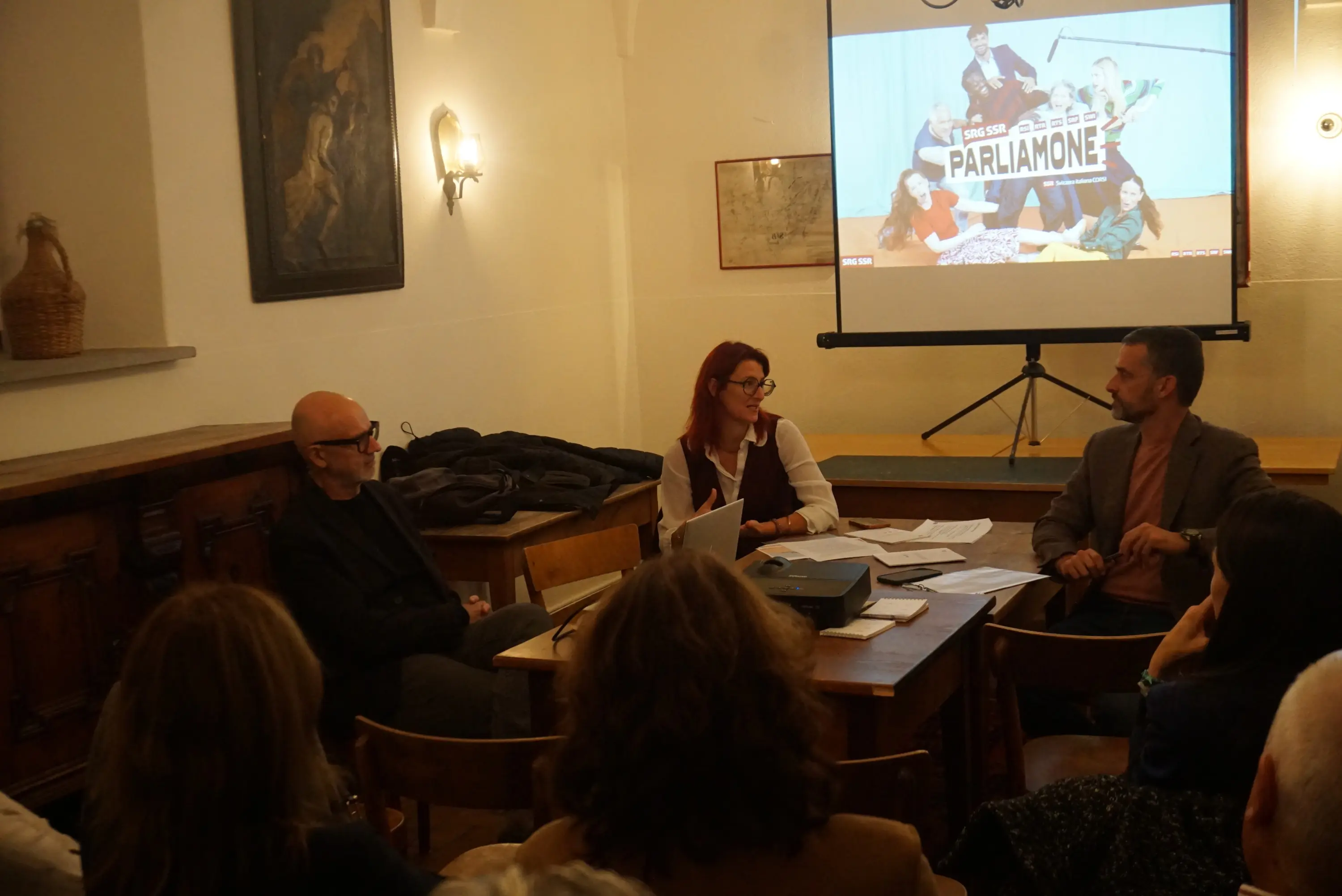 Incontro di discussione con tre relatori seduti attorno a un tavolo. Sullo sfondo, una proiezione con la scritta "Parliamone". I partecipanti sembrano coinvolti nella conversazione.