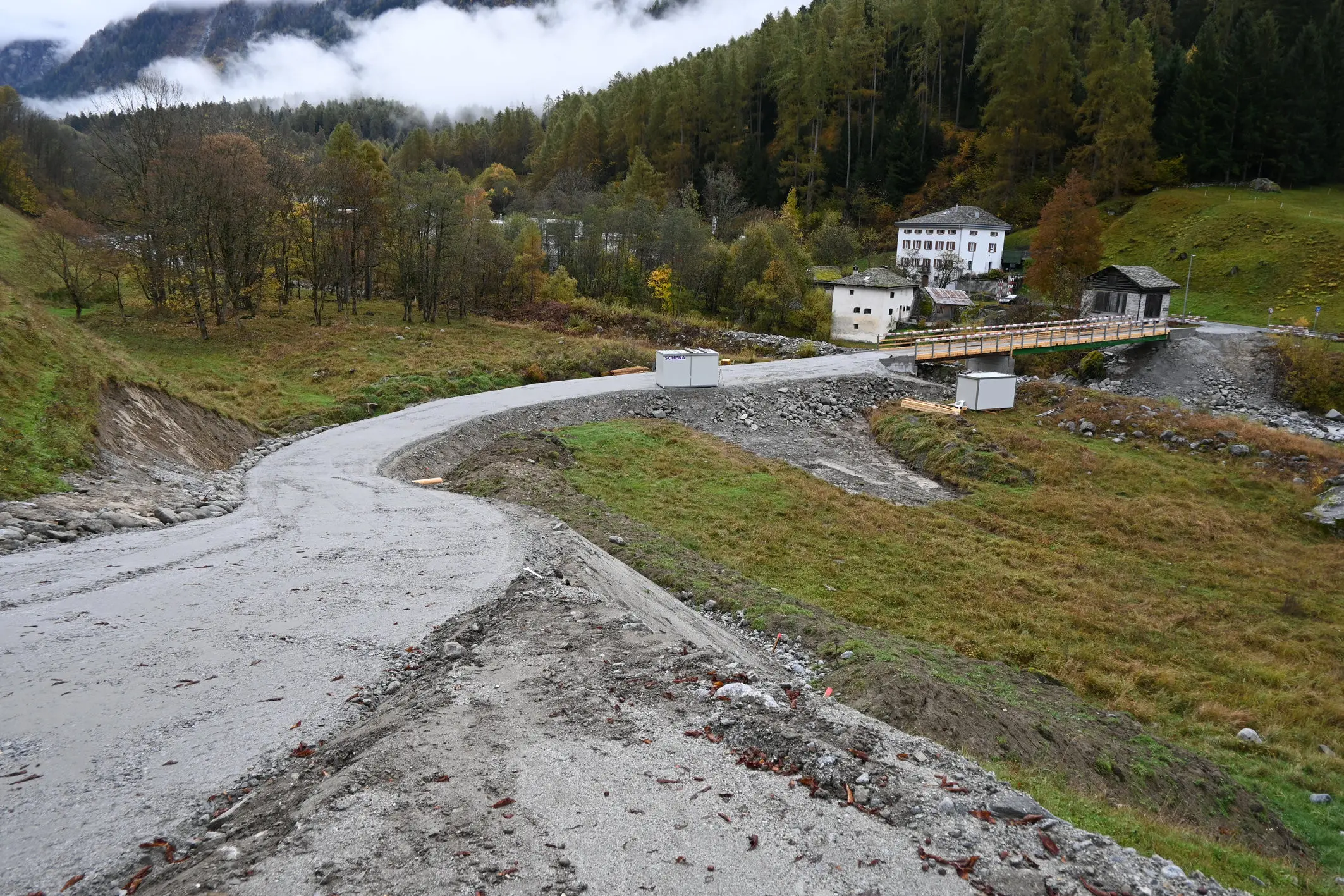 Una strada sinuosa in un paesaggio montano, con alberi e nuvole sullo sfondo. A destra, si intravedono alcune costruzioni, tra cui una casa e un ponte. Il terreno è in parte spoglio e presenta macchie di ghiaia.